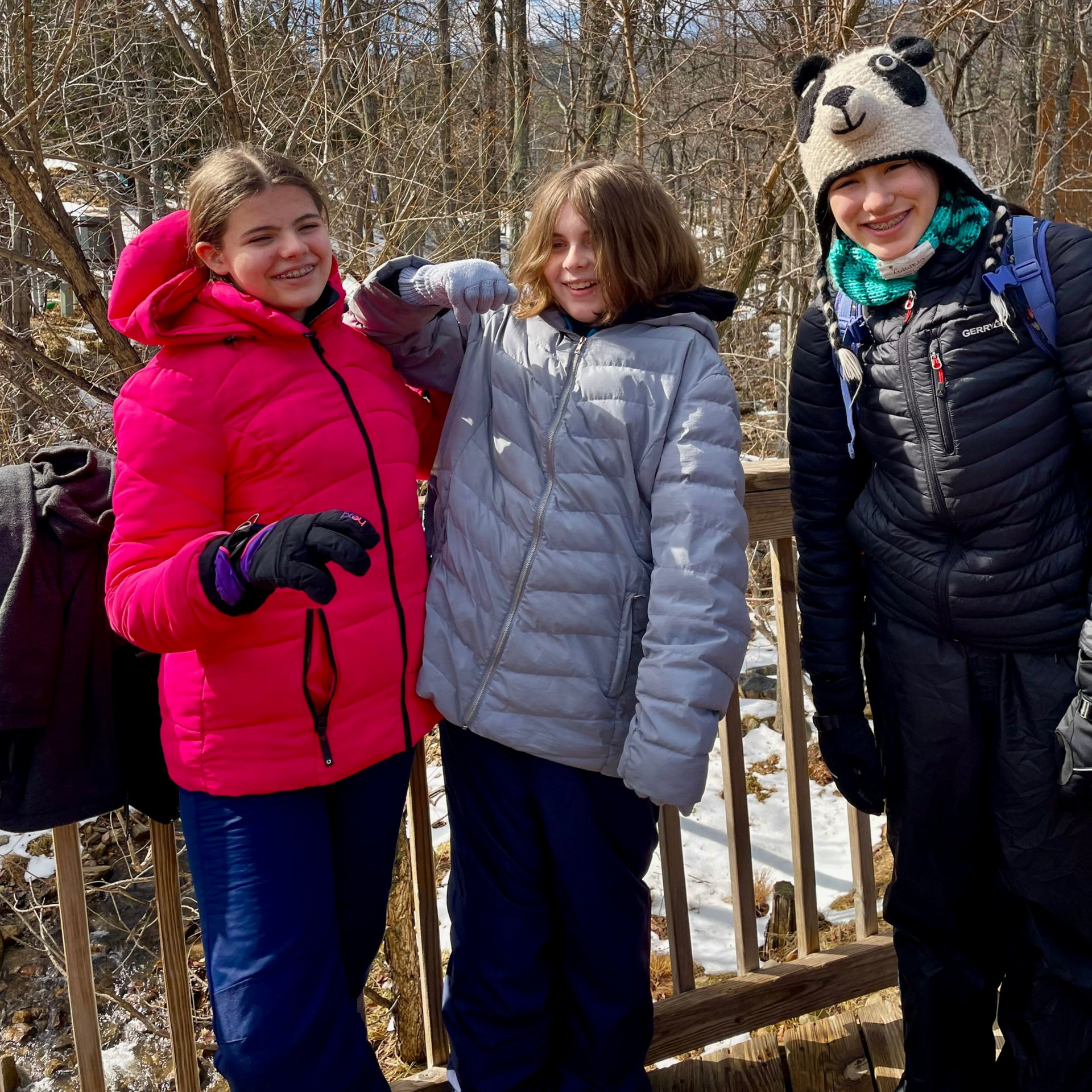 Three people in winter jackets stand on a wooden deck with snow, one in a panda hat.