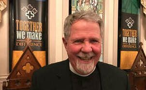 Man with gray beard smiles, wearing a black collar in a church with banner 