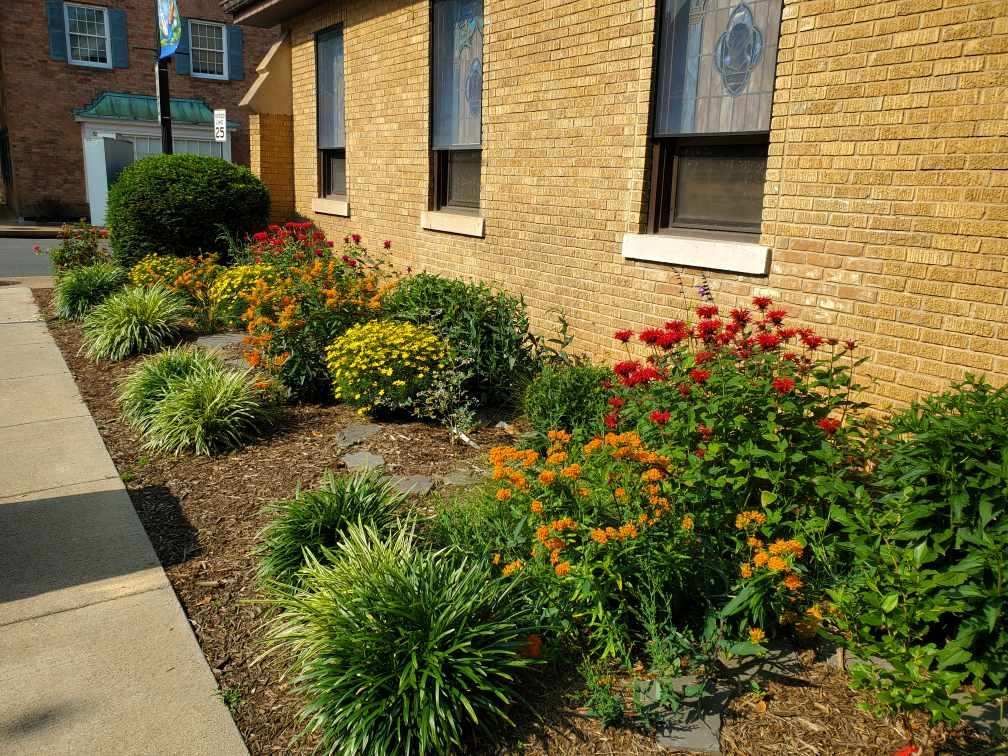 Flower bed with various flowers and shrubs next to a brick building.