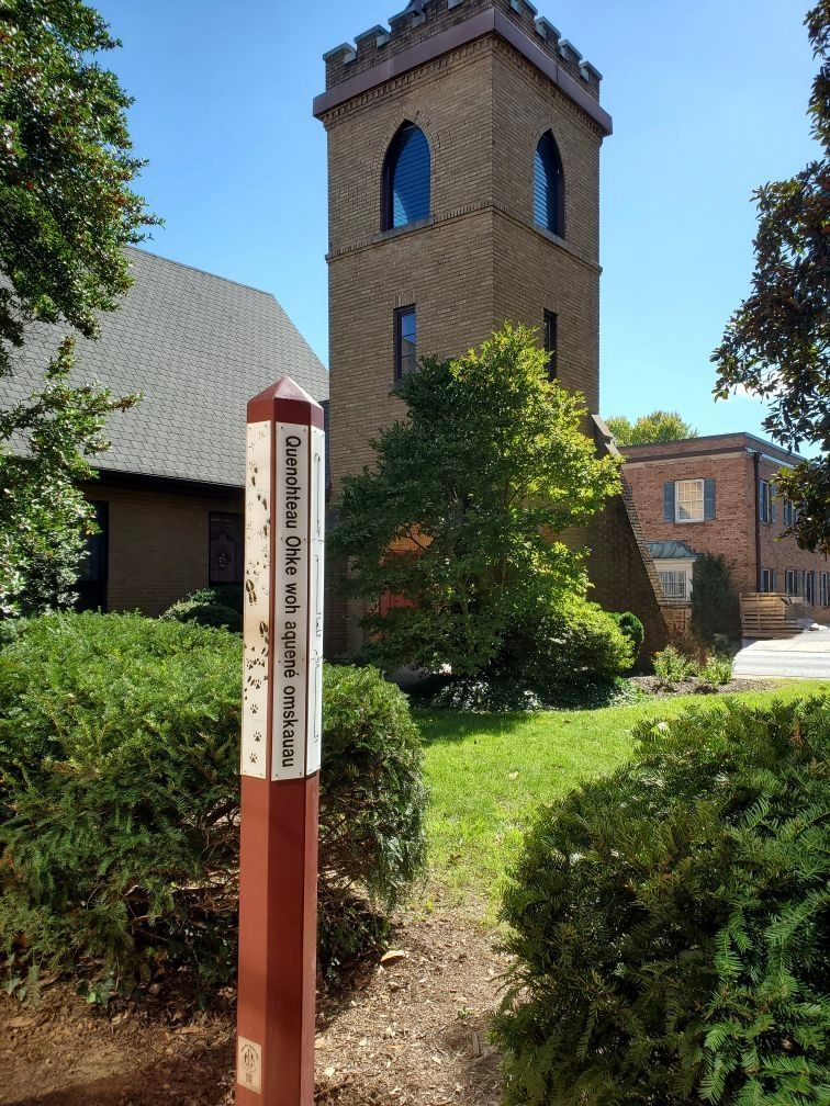 Tall brick tower with arched windows, sign in foreground, green lawn and bushes, blue sky.