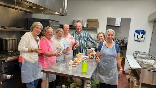 Group of people in a kitchen, smiling and holding food items, near a stainless steel table with food.