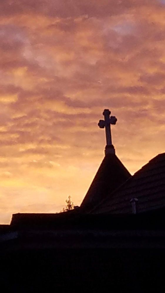 Silhouette of a church roof and cross against a colorful sunset sky.