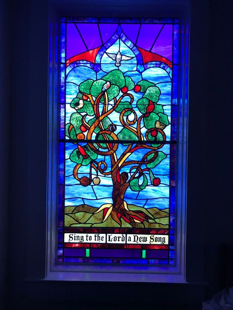 Stained glass window depicting a tree with musical notes as fruit, a dove, and text 