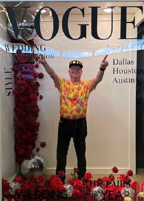 Man posing inside a Vogue wedding display, surrounded by red flowers and city names on the wall
