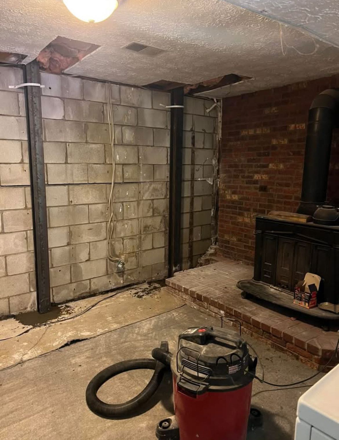 Basement renovation with cinder block wall, steel beams, brick fireplace, and wet/dry vacuum on concrete floor.