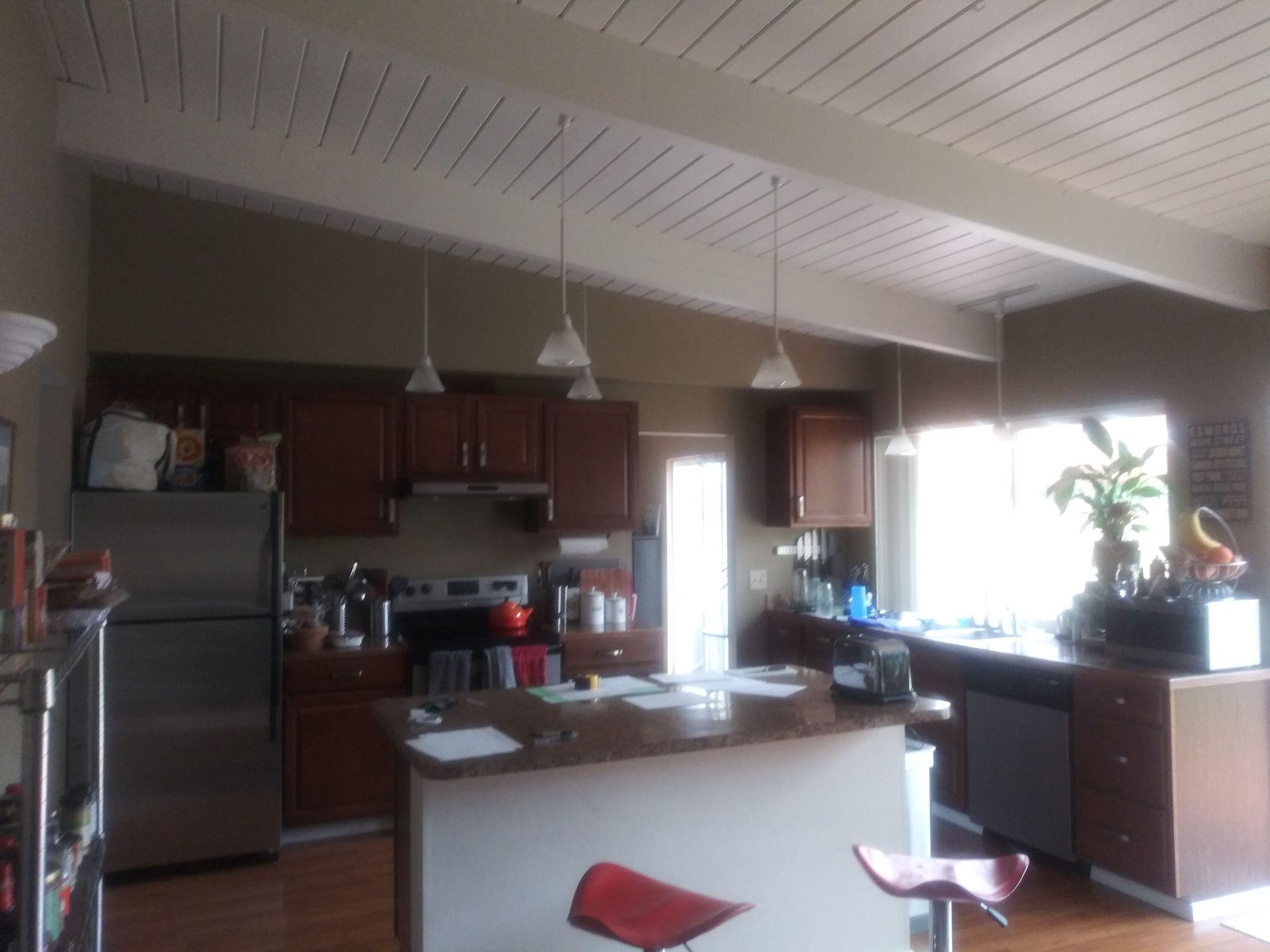 Modern kitchen with stainless fridge, wood cabinets, island, pendant lights, and bright window by sink.