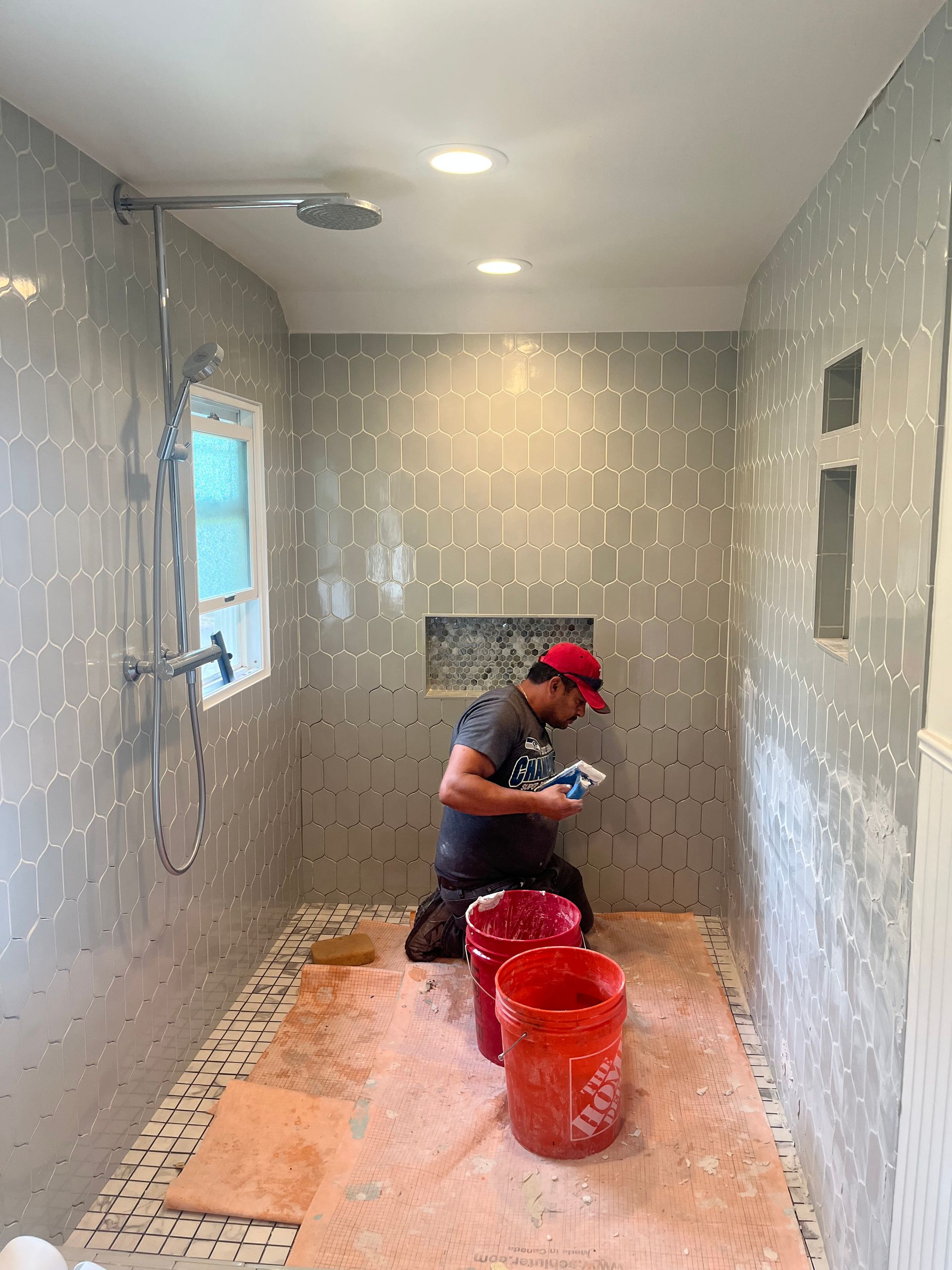 Person kneels in a tiled shower, cleaning with a sponge beside red buckets and a window.