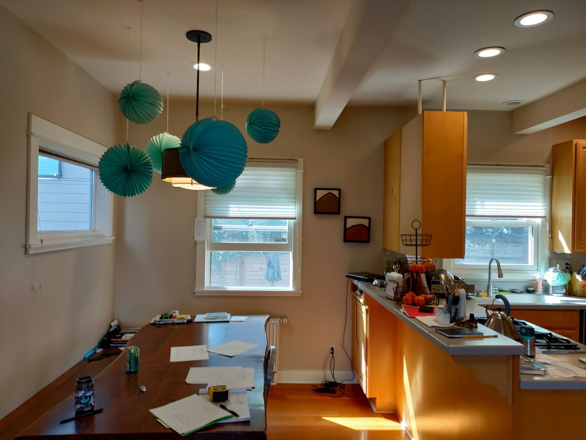 Bright kitchen with wooden table, orange cabinets, and teal hanging paper lanterns by two windows.