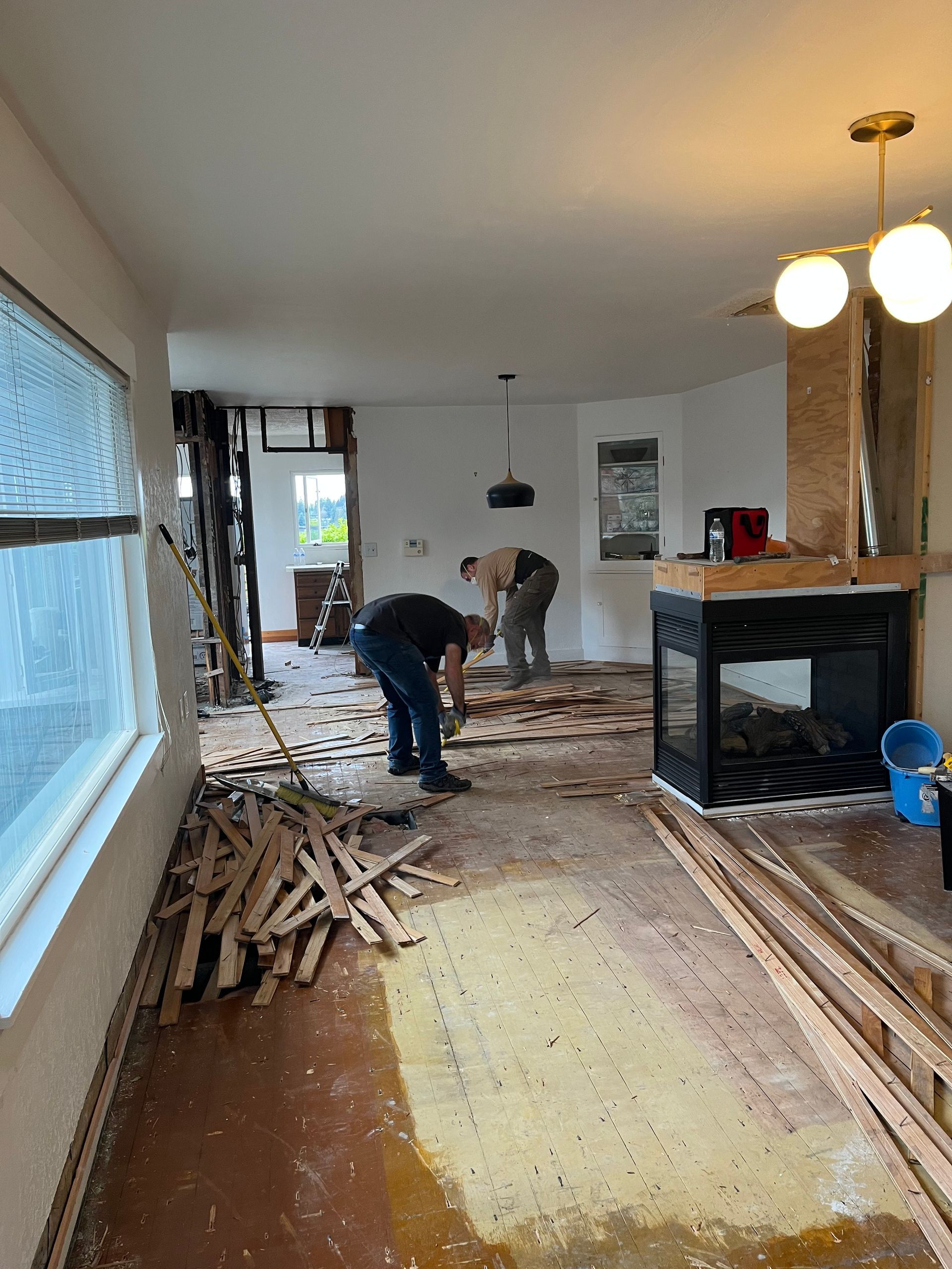 Two people clearing debris in a damaged room with wood planks, broken flooring, and a hanging light fixture.