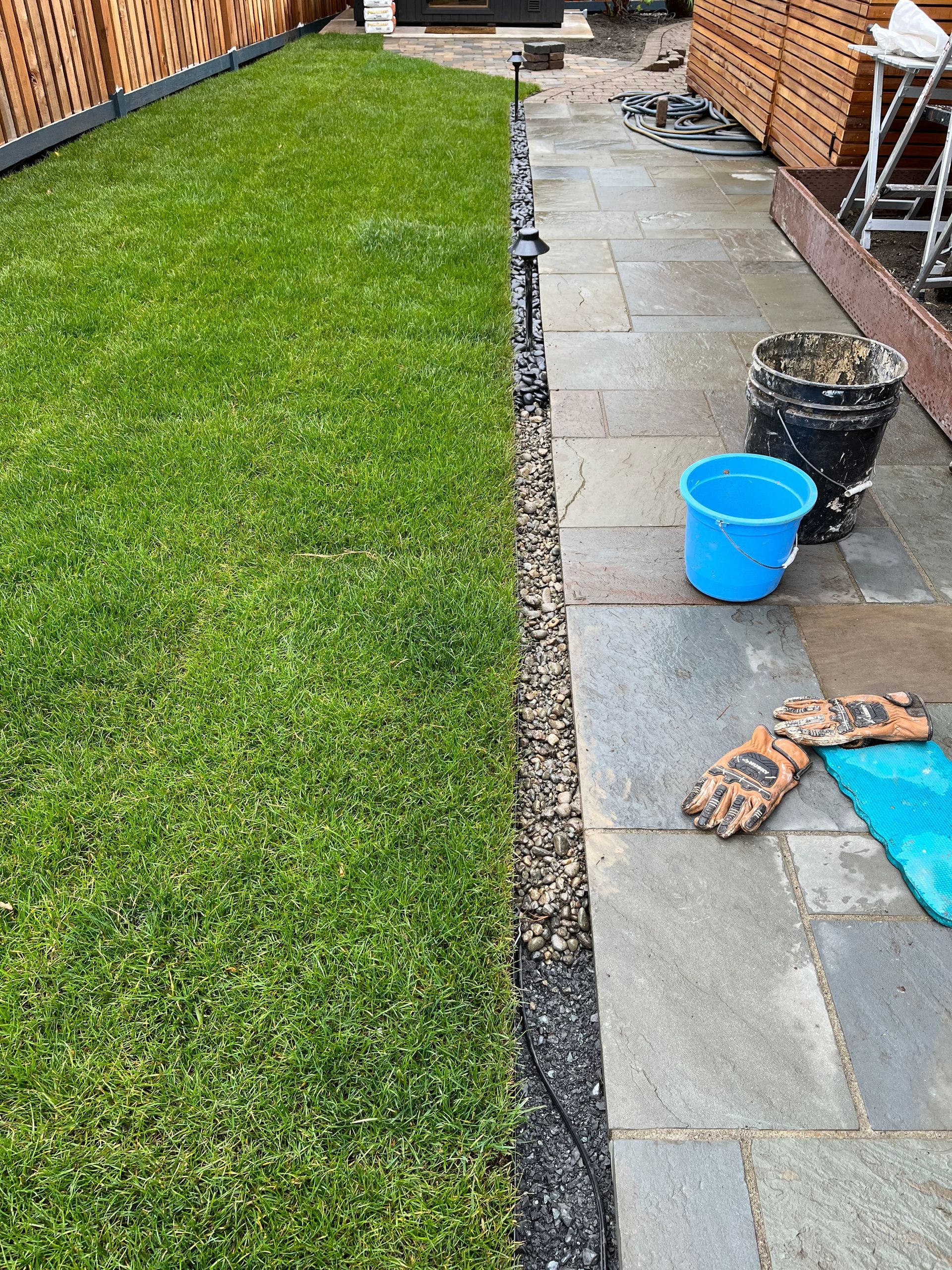 Backyard patio with a lawn on the left, stone path on the right, and a blue bucket and gloves near the foreground
