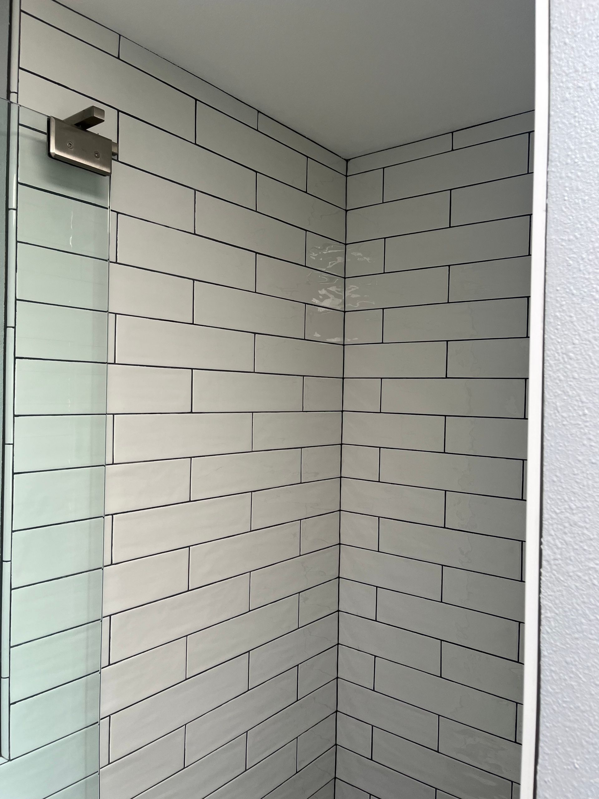 Tiled shower corner with white tiles, glass door edge, and a wall-mounted showerhead from above