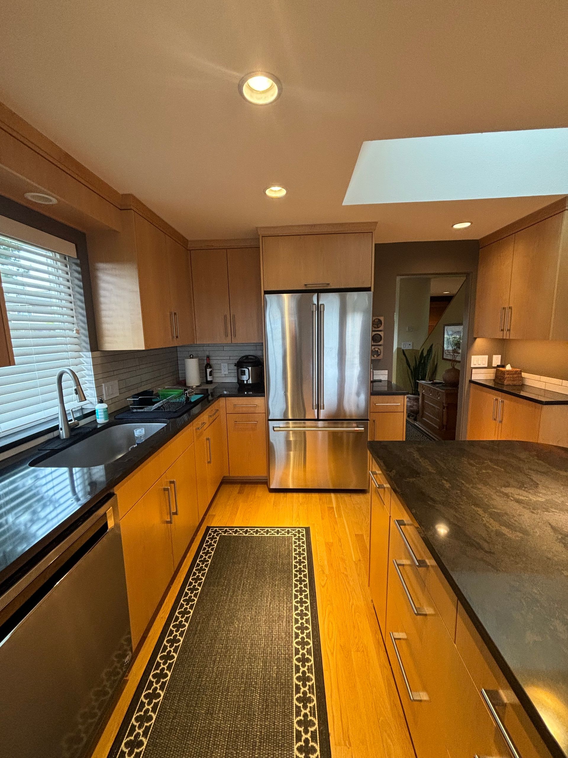 Modern kitchen with stainless steel appliances, wooden cabinets, black countertops, and a skylight.