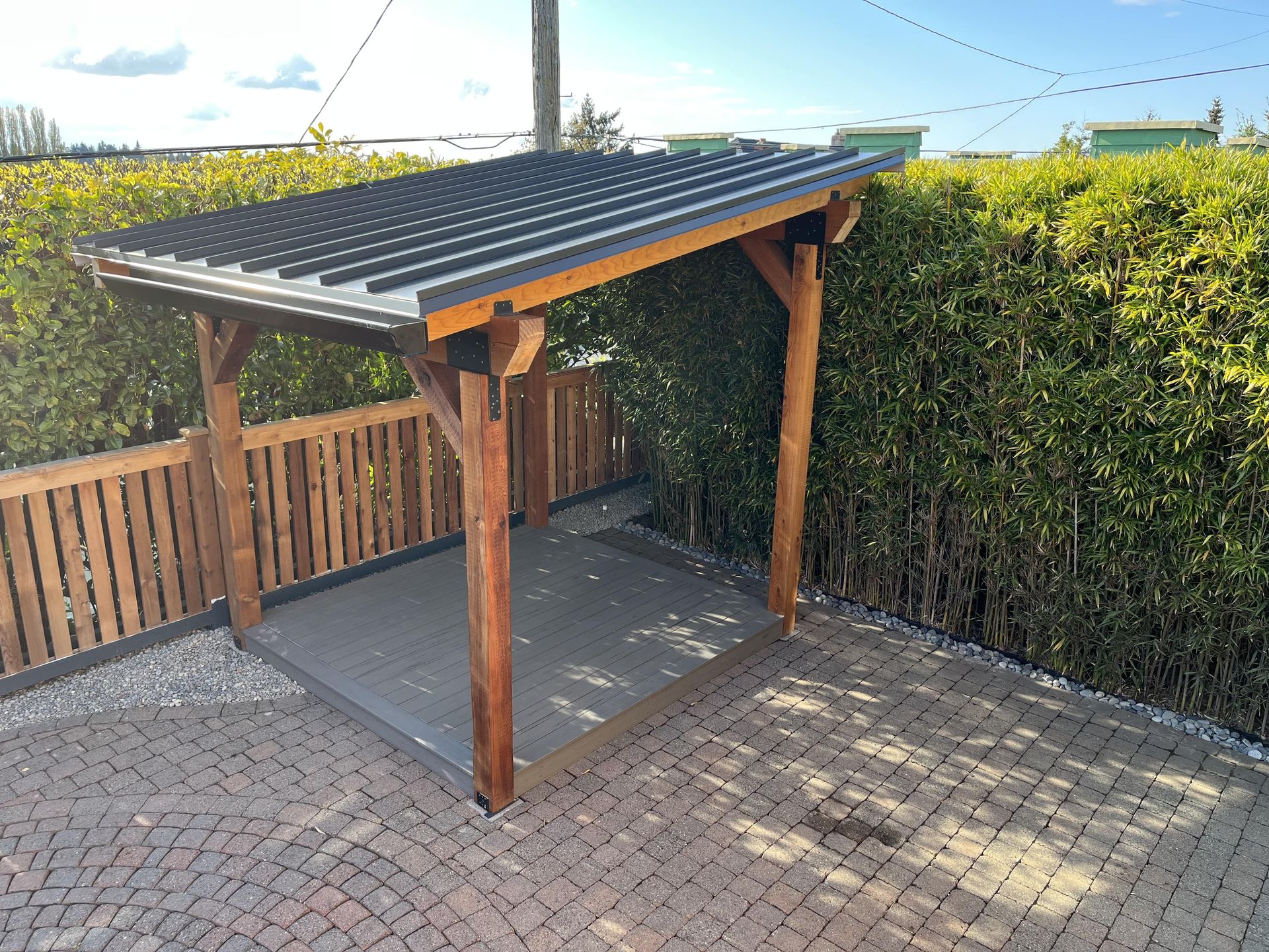 Small wooden carport with a corrugated roof on a paved driveway, beside a hedge and fence.