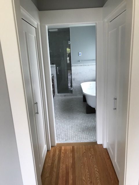 Narrow hallway with wood floor leading to a bathroom with gray tile and a white bathtub