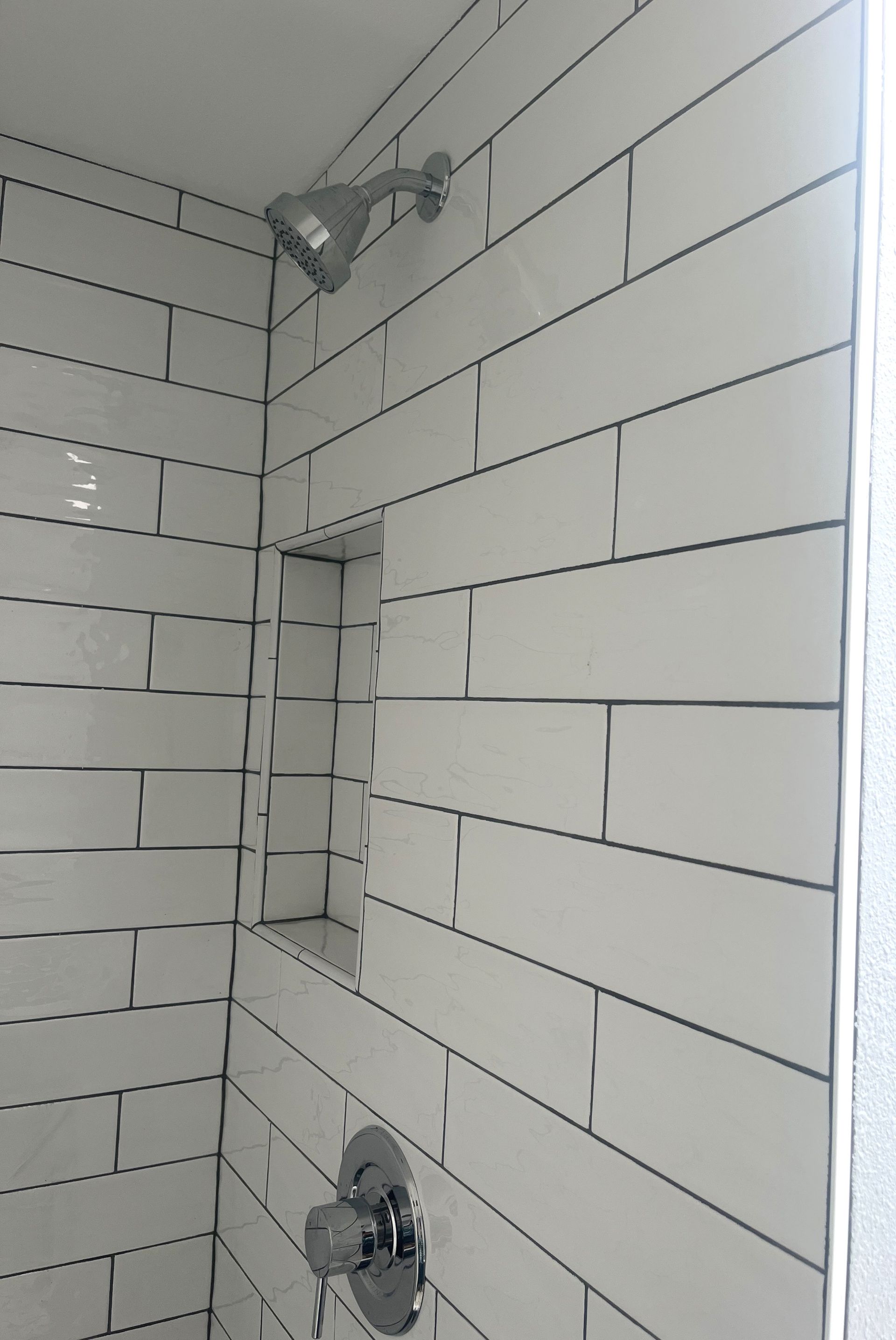 White tiled bathroom wall with shower head, recessed shelf, and silver faucet.