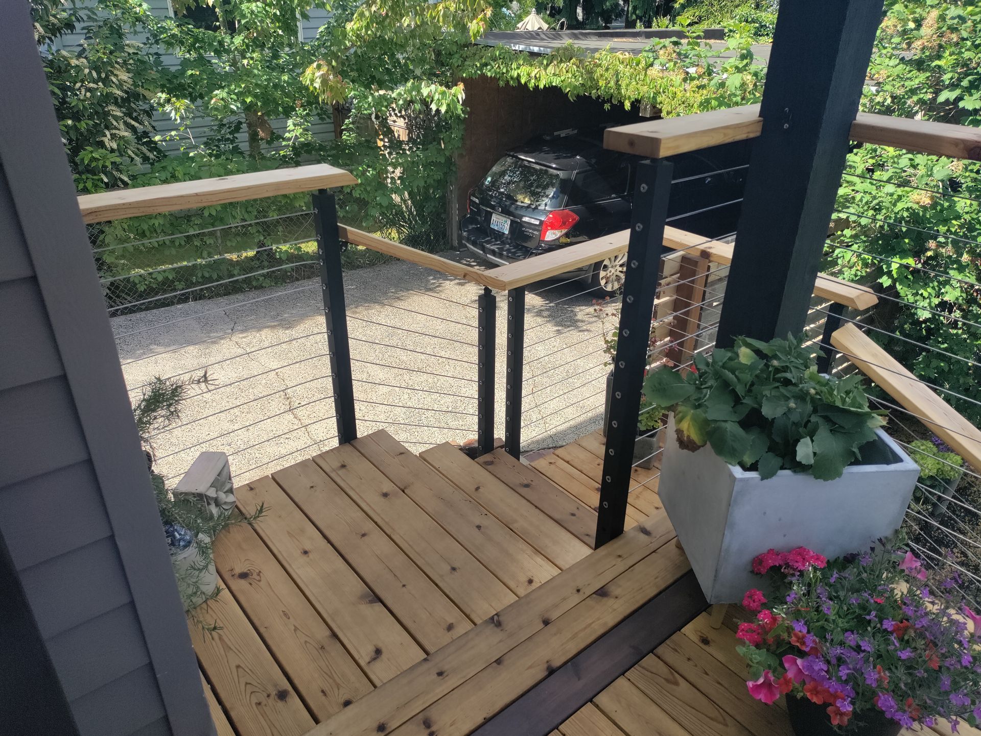 Small wooden porch with grill, potted flowers, and a garden view through the railing.
