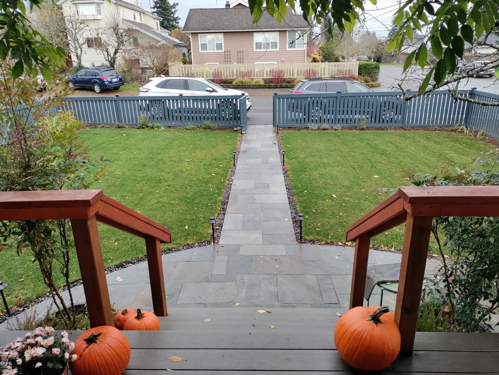 Front porch with pumpkins, stone walkway, and a yard leading to a blue fence and houses across the street.