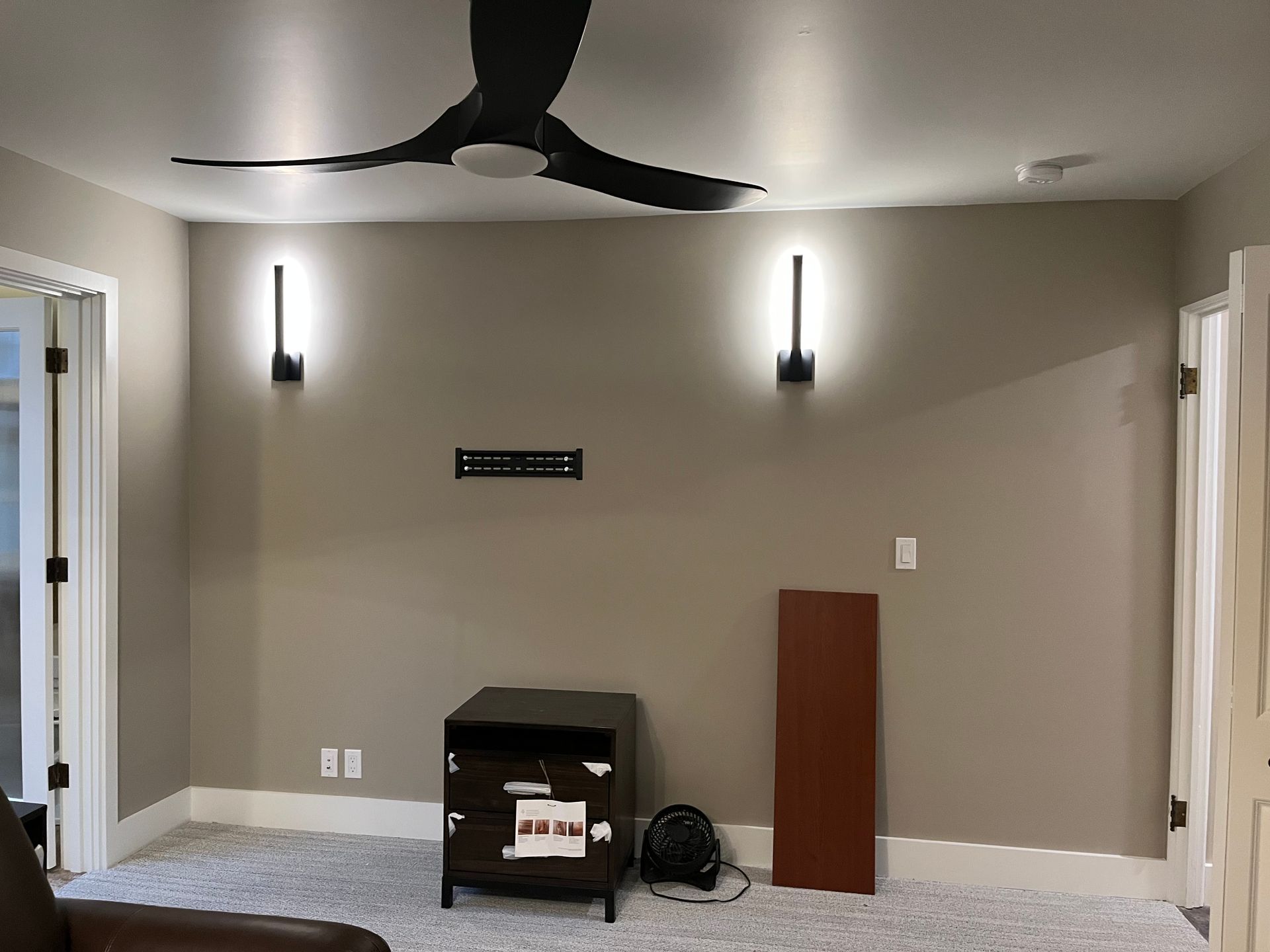 Empty room with beige walls, ceiling fan, wall sconces, and a small fireplace or heater against the back wall