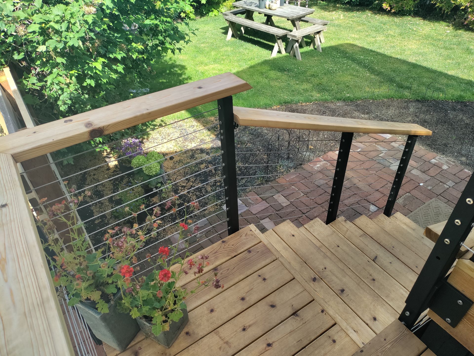 Wooden deck stairs with cable railing leading down to a brick patio and green backyard garden.