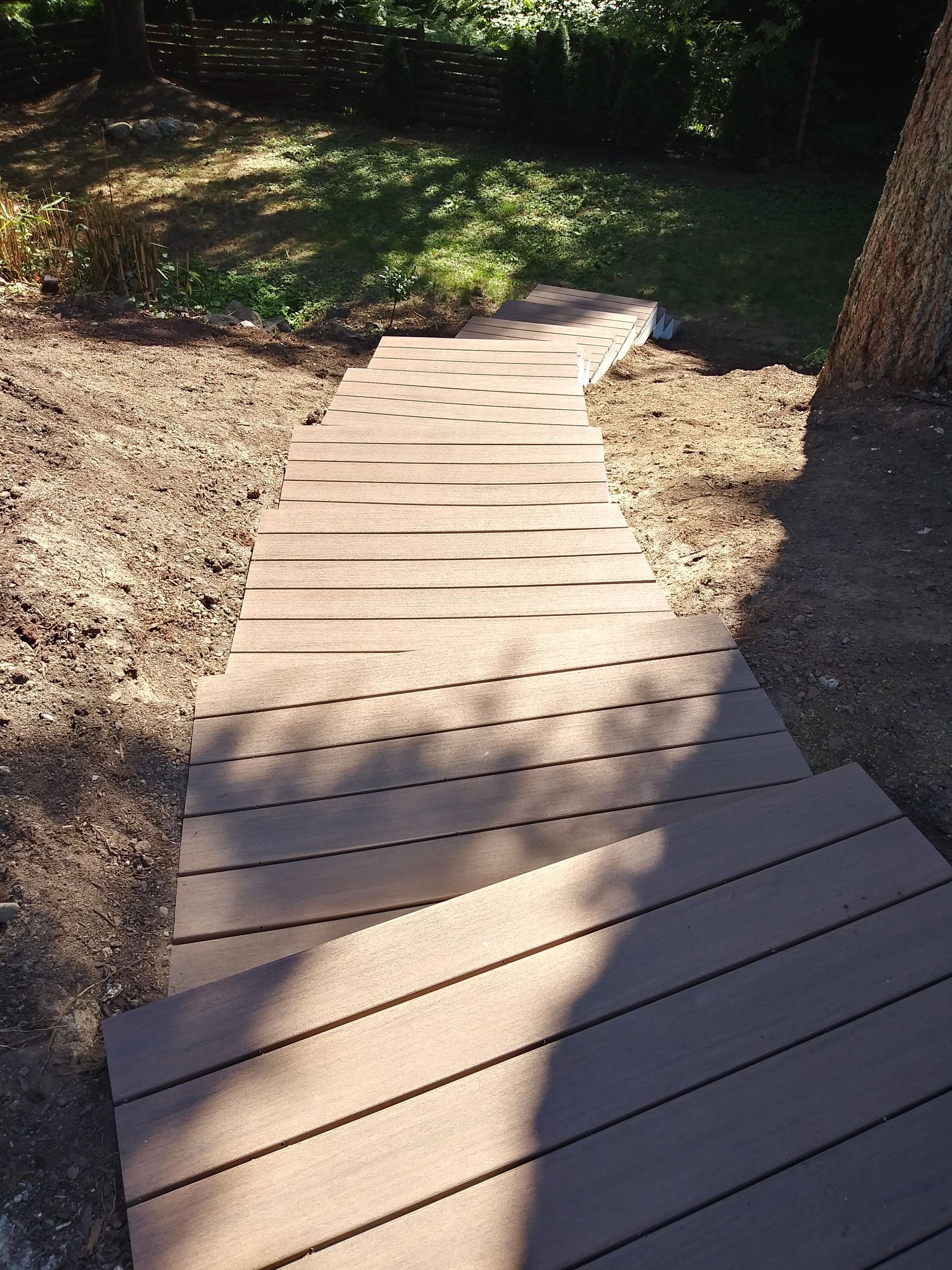 Wooden boardwalk path through a shaded garden, curving past trees and dirt ground.