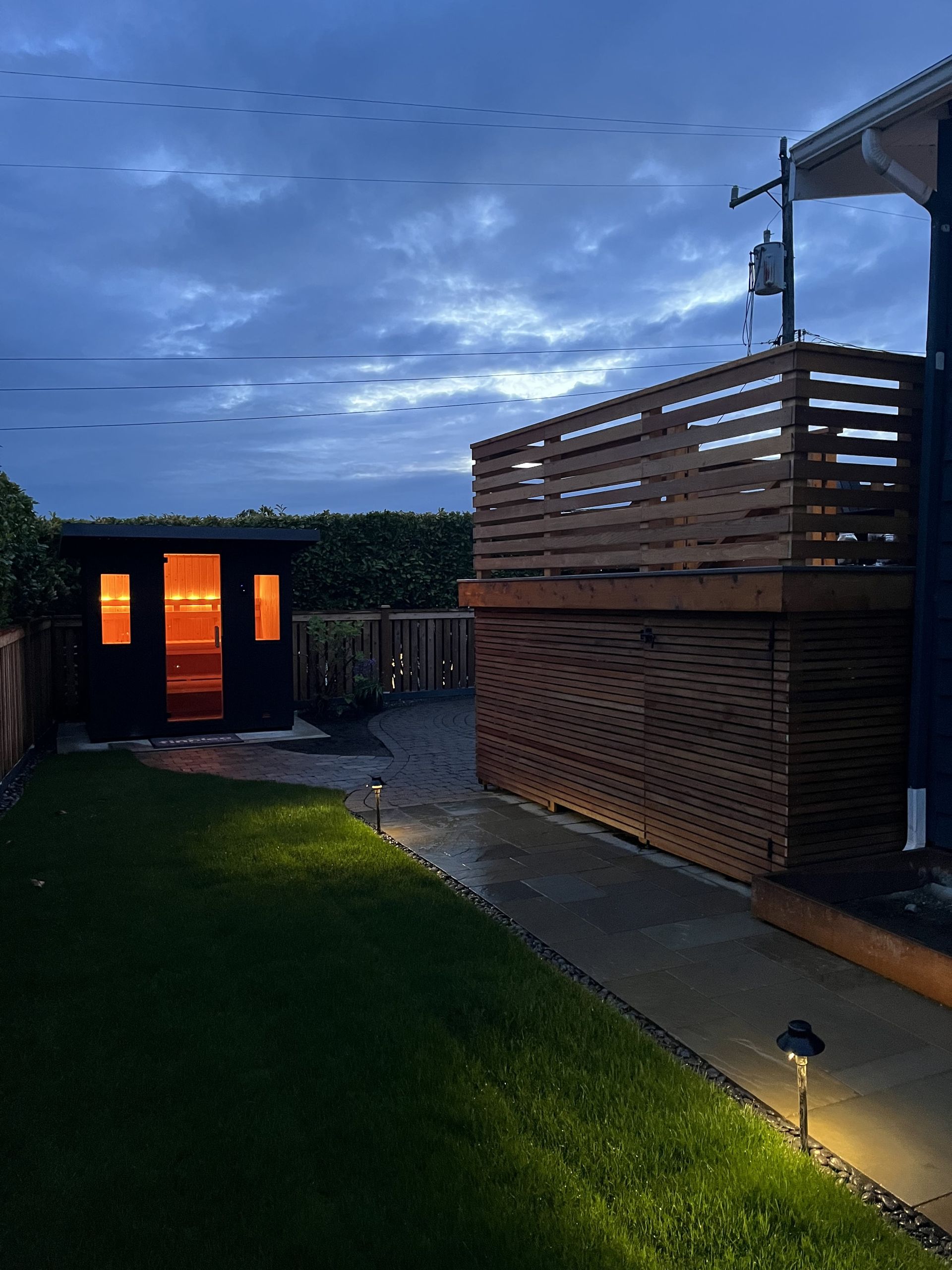 Dusk backyard with lit grass path, wooden deck, and small building glowing orange in the distance