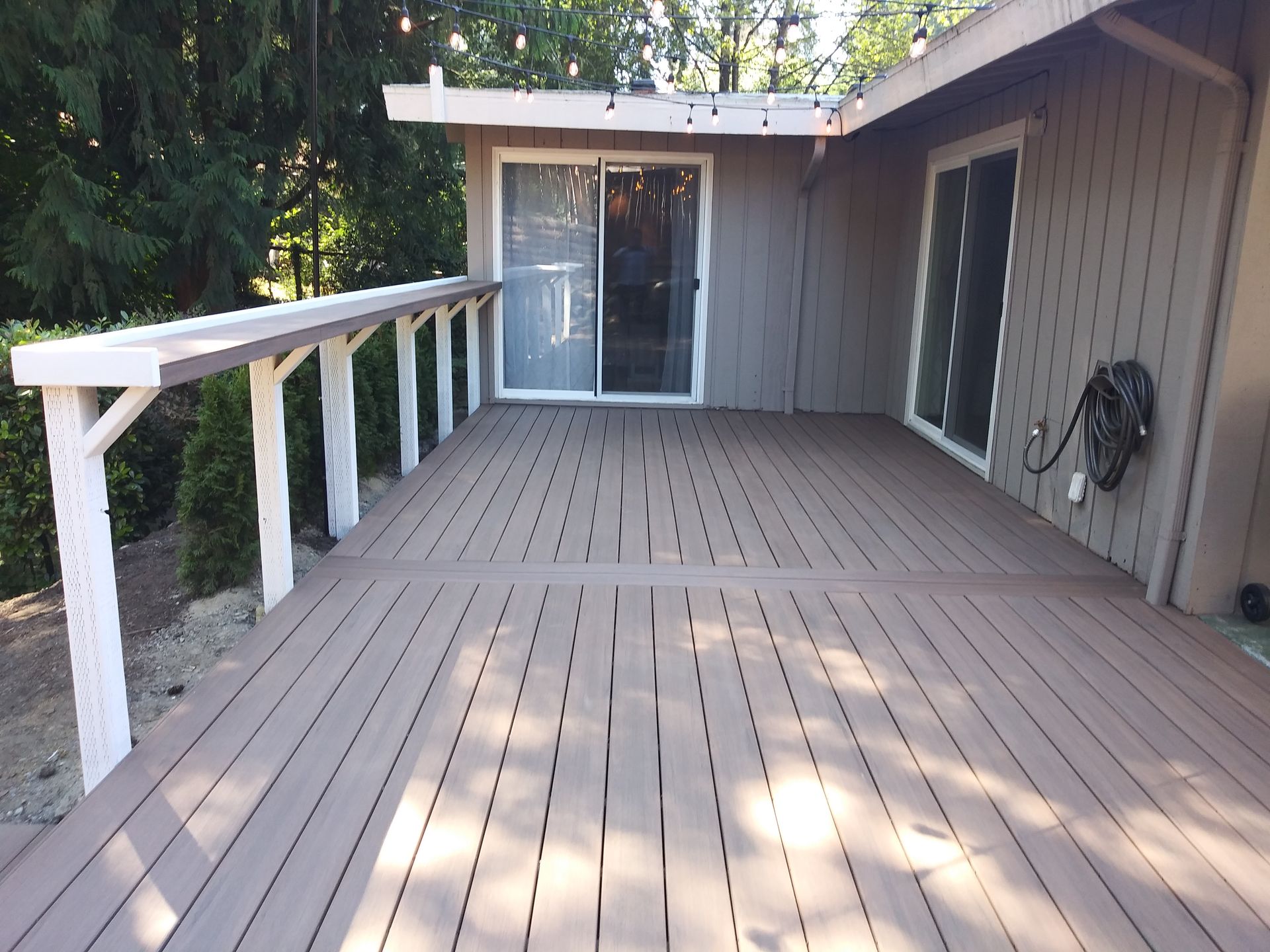 Wooden deck with white railing, leading to a sliding glass door beside a house and trees.