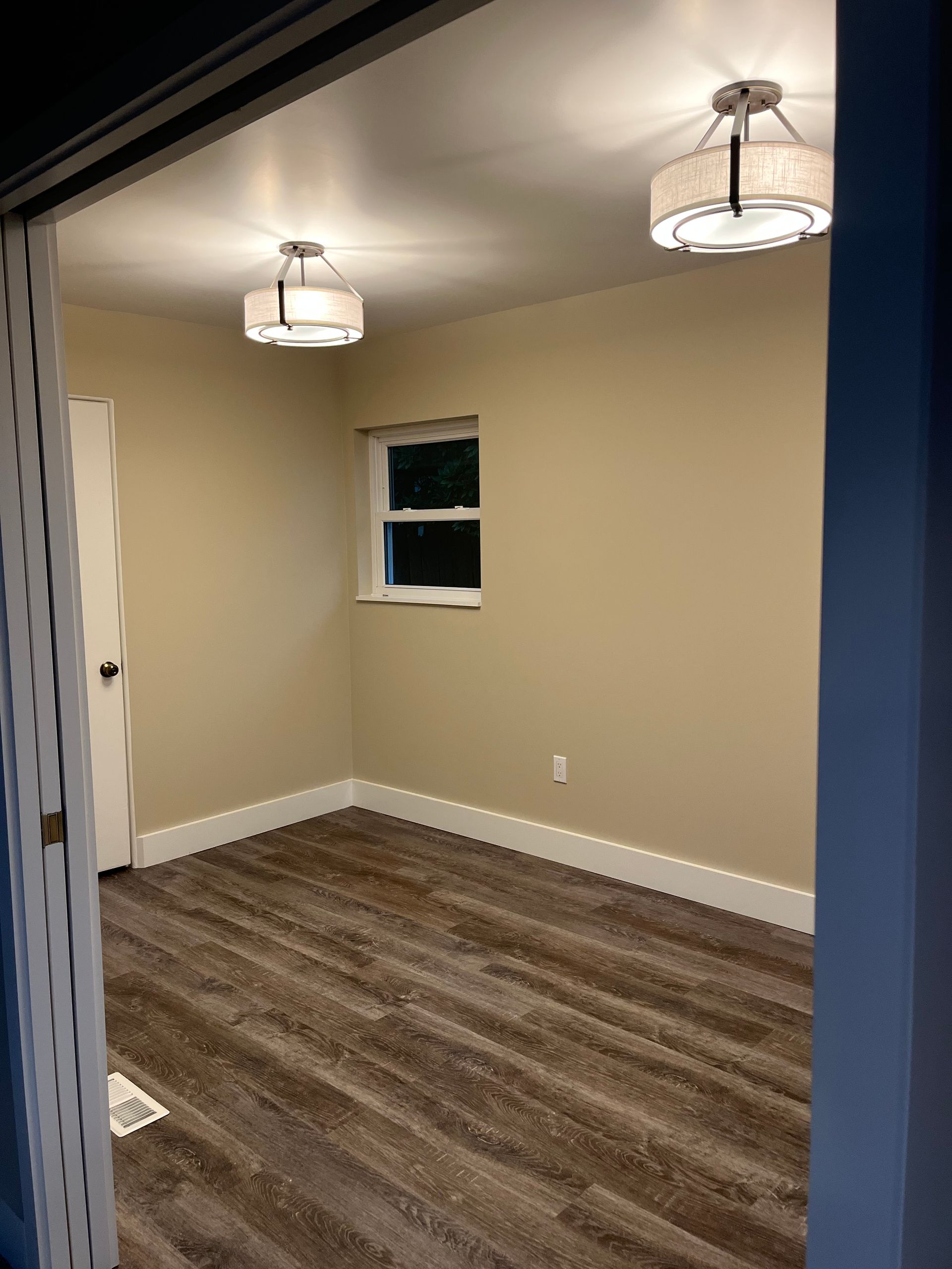 Empty beige room with wood flooring, two ceiling lights, and a small window.