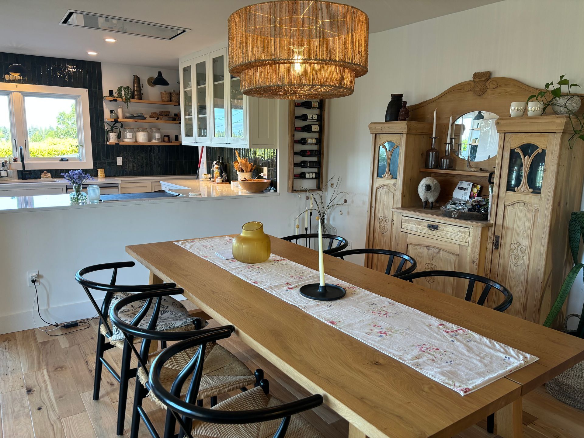 Modern dining room with wooden table, black chairs, wicker pendant light, and white kitchen in background