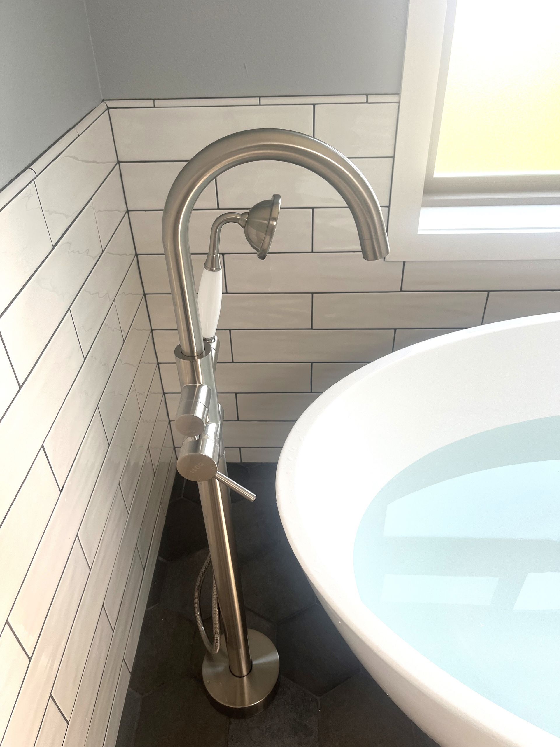 Tall chrome bathroom faucet beside a white sink against white tiled walls and a window.