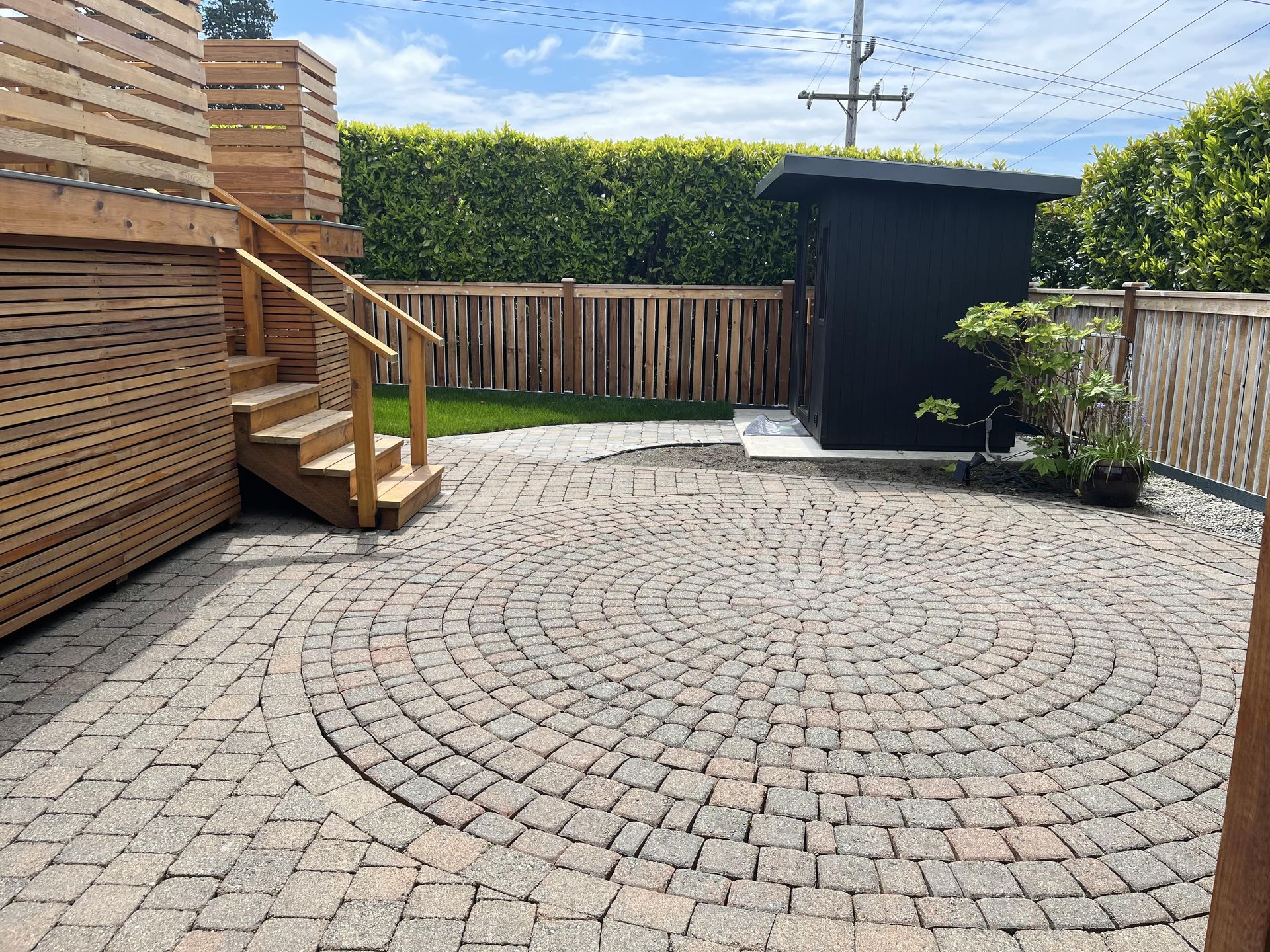 Backyard patio with circular cobblestone paving, wooden deck stairs, fencing, and a small dark shed