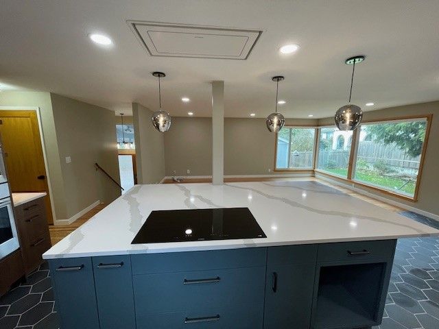Modern kitchen with large white island, black cooktop, pendant lights, and big windows overlooking greenery