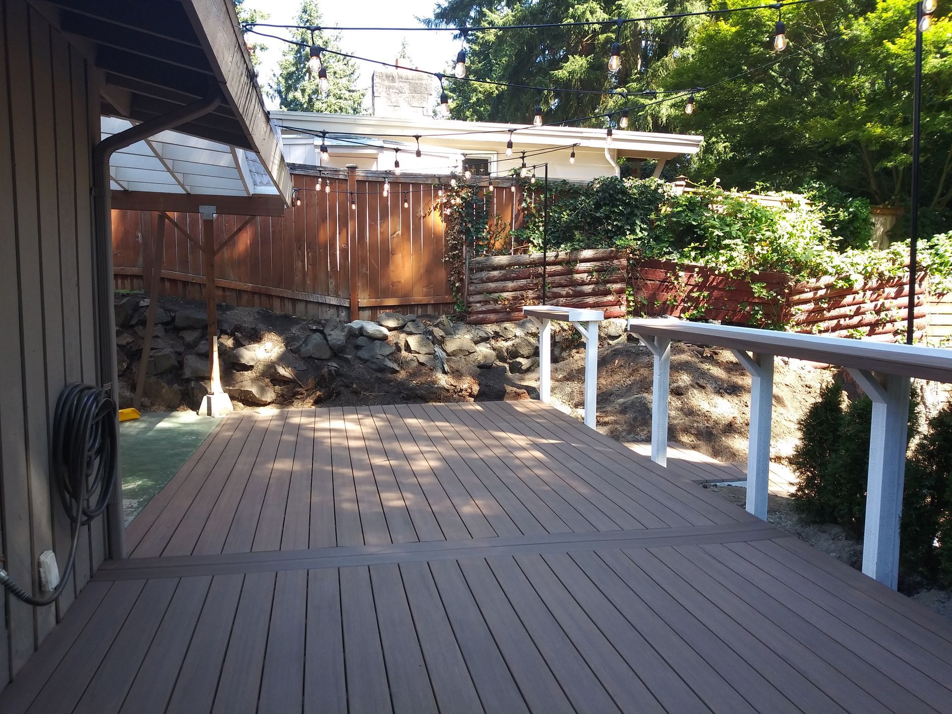 Wooden deck walkway beside a house, with a dog resting near a stone wall and trees in the background