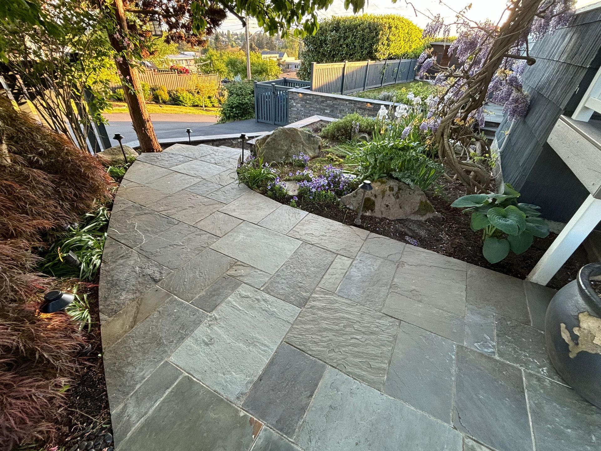 Curved stone pathway through a garden with trees, shrubs, and a low stone wall