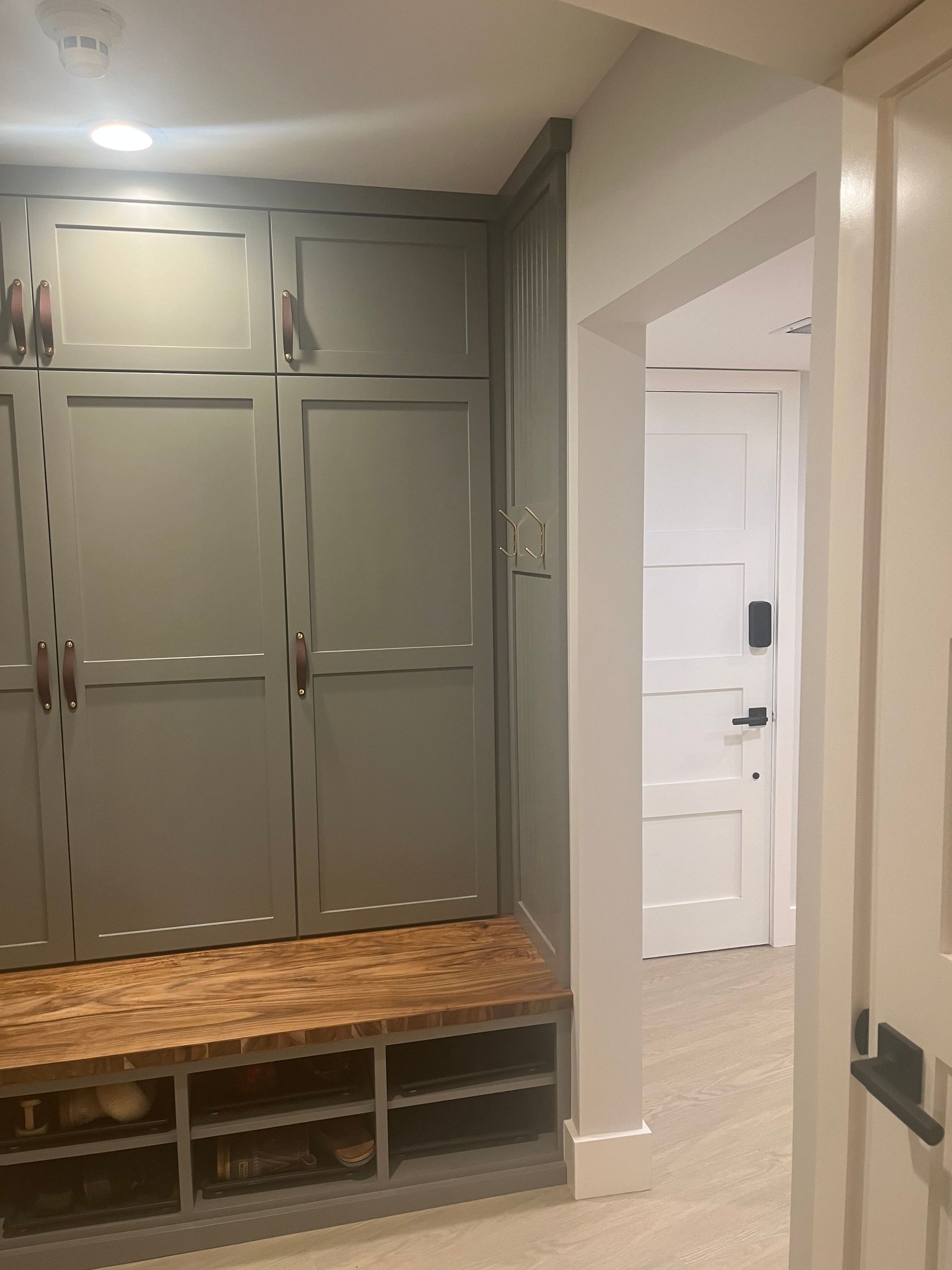 Mudroom with gray built-in cabinets, wooden bench, cubbies, and a white door in the hallway.