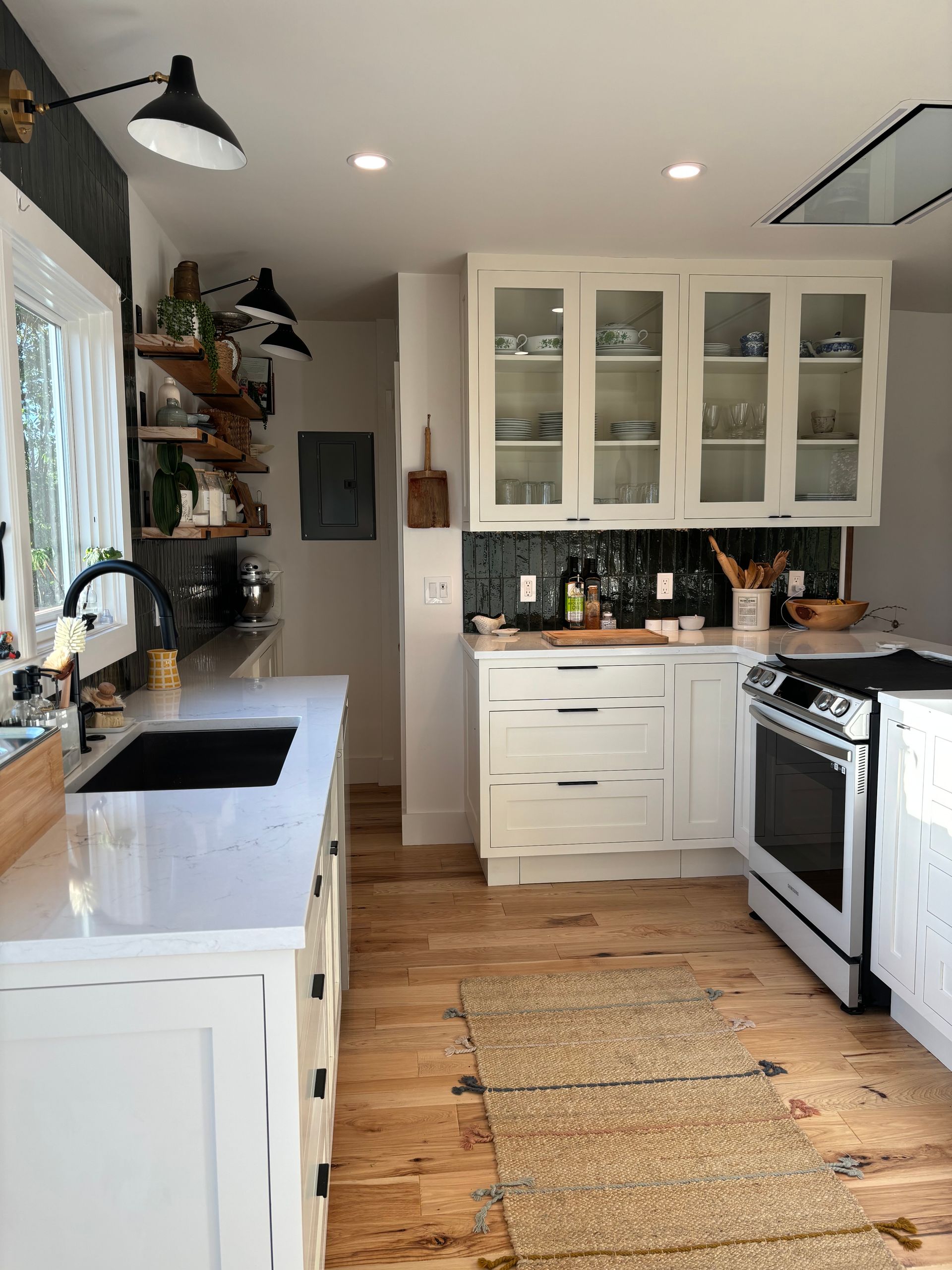 Bright white kitchen with island, glass-front cabinets, black stove, woven runner, and shelves by a window.