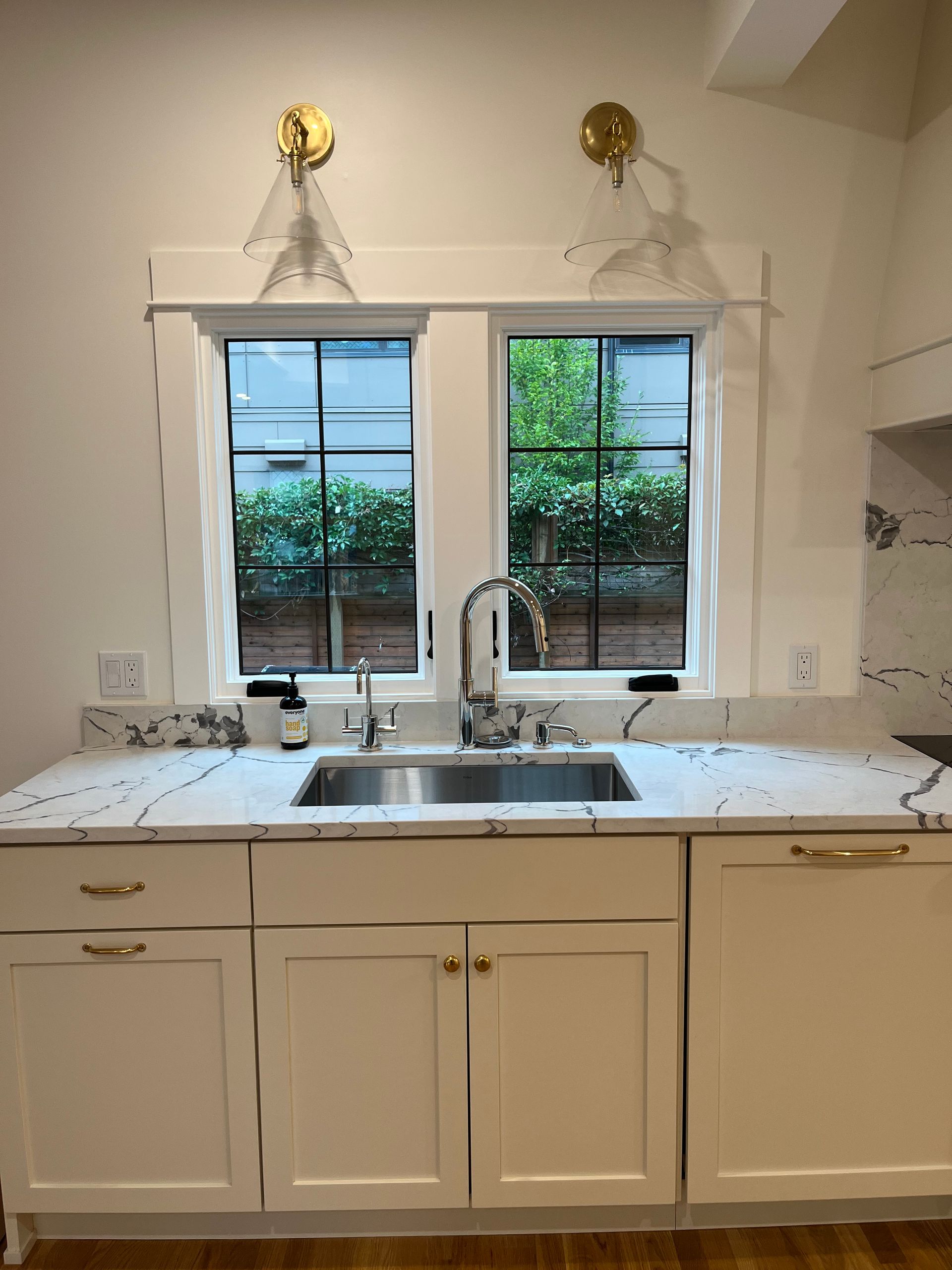 Bright kitchen sink under two windows with white cabinets and brass wall lights