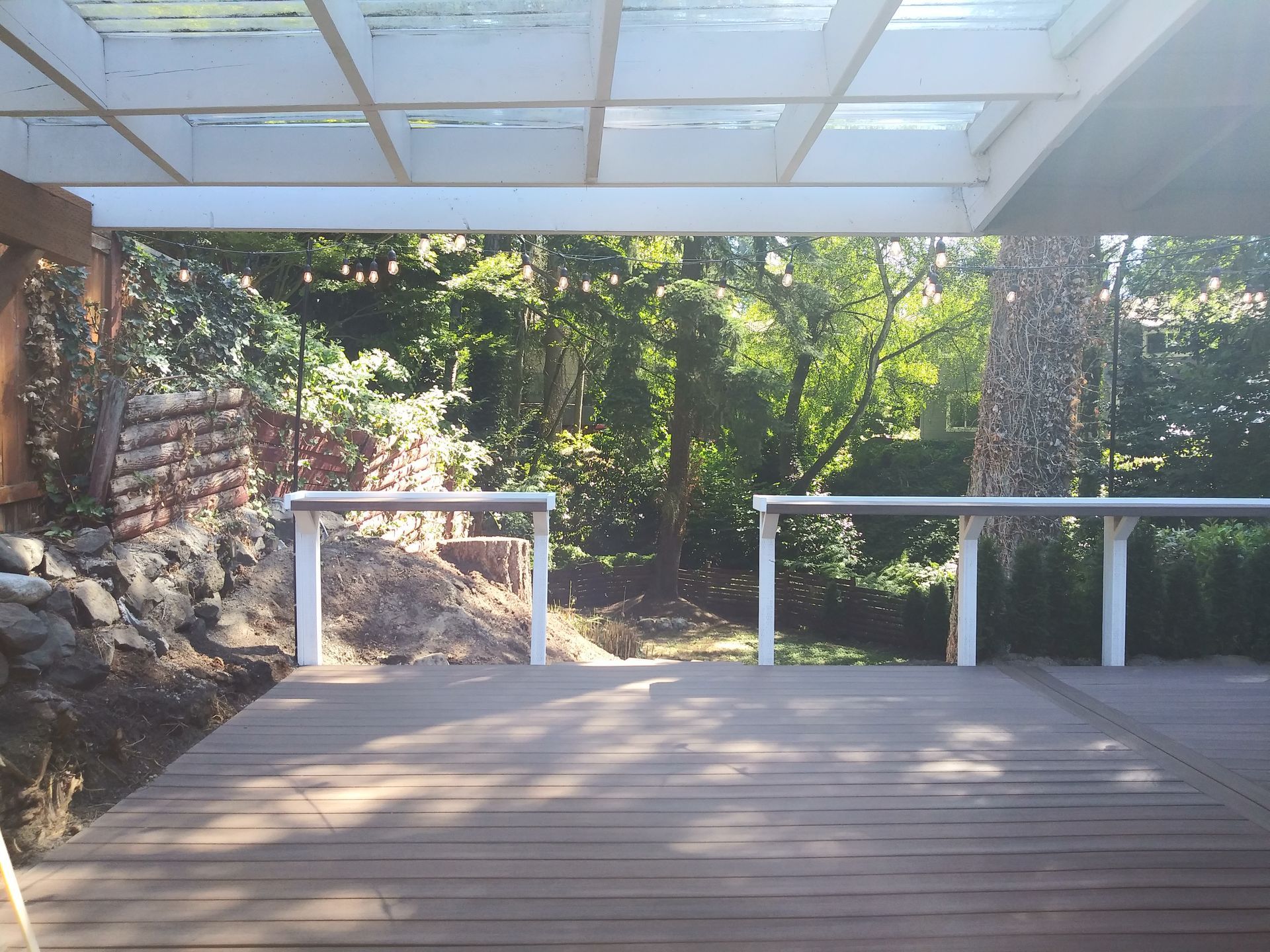 Covered wooden deck with white railings overlooking a sunlit garden and stone wall.
