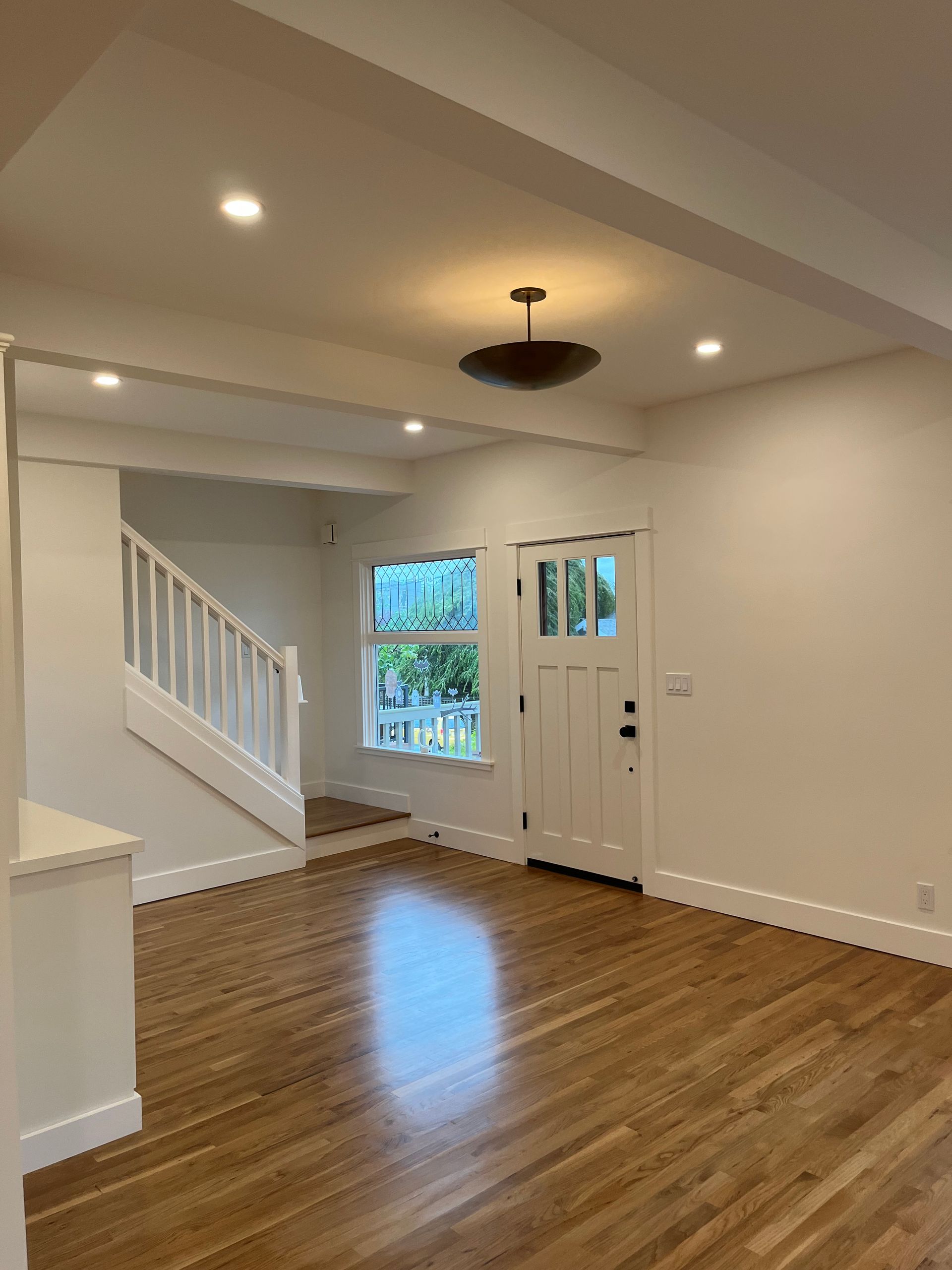 Empty living room with hardwood floors, white walls, recessed lights, staircase, and front door.