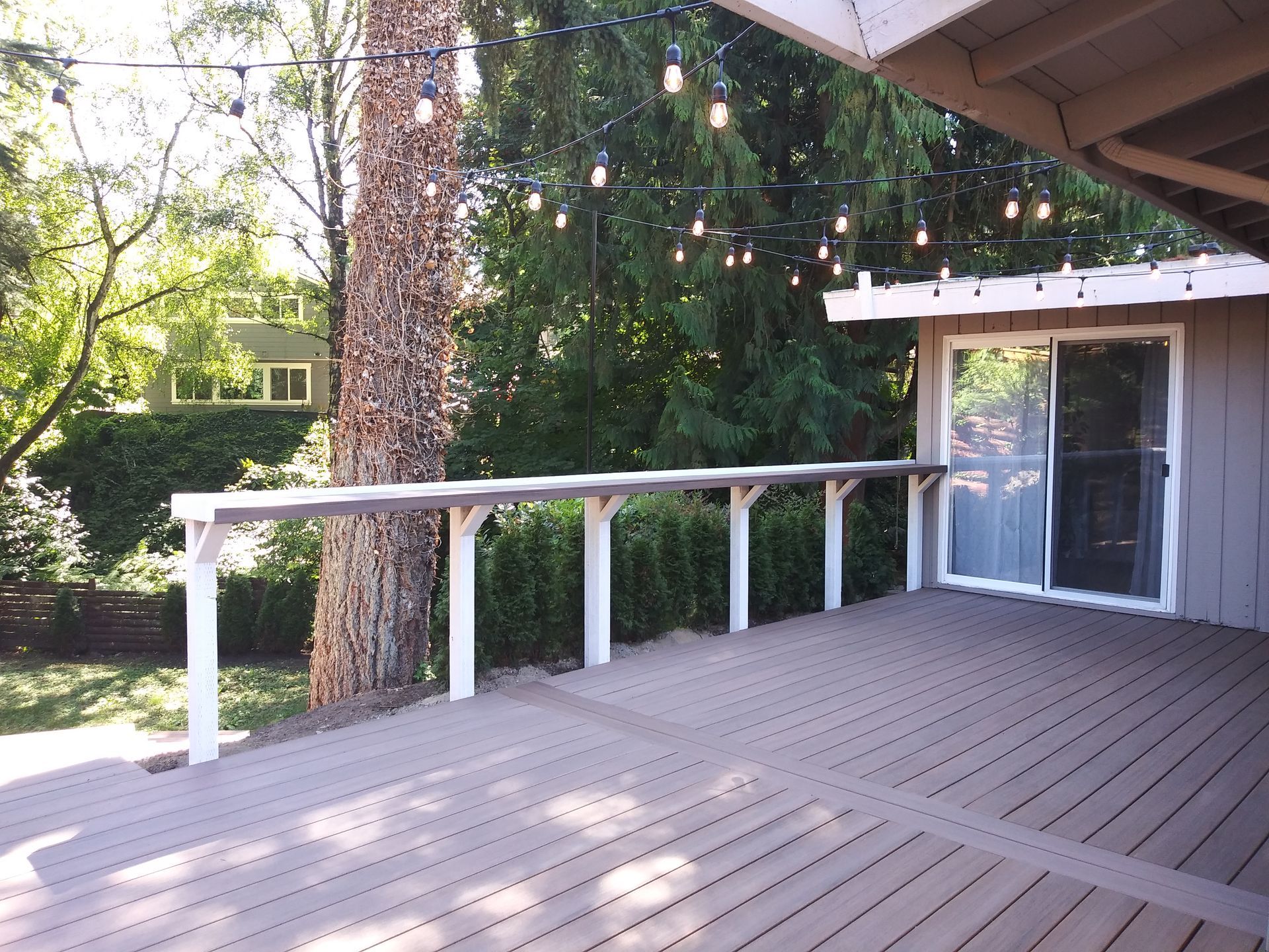 Wooden deck with white railing and string lights beside a house and large tree.