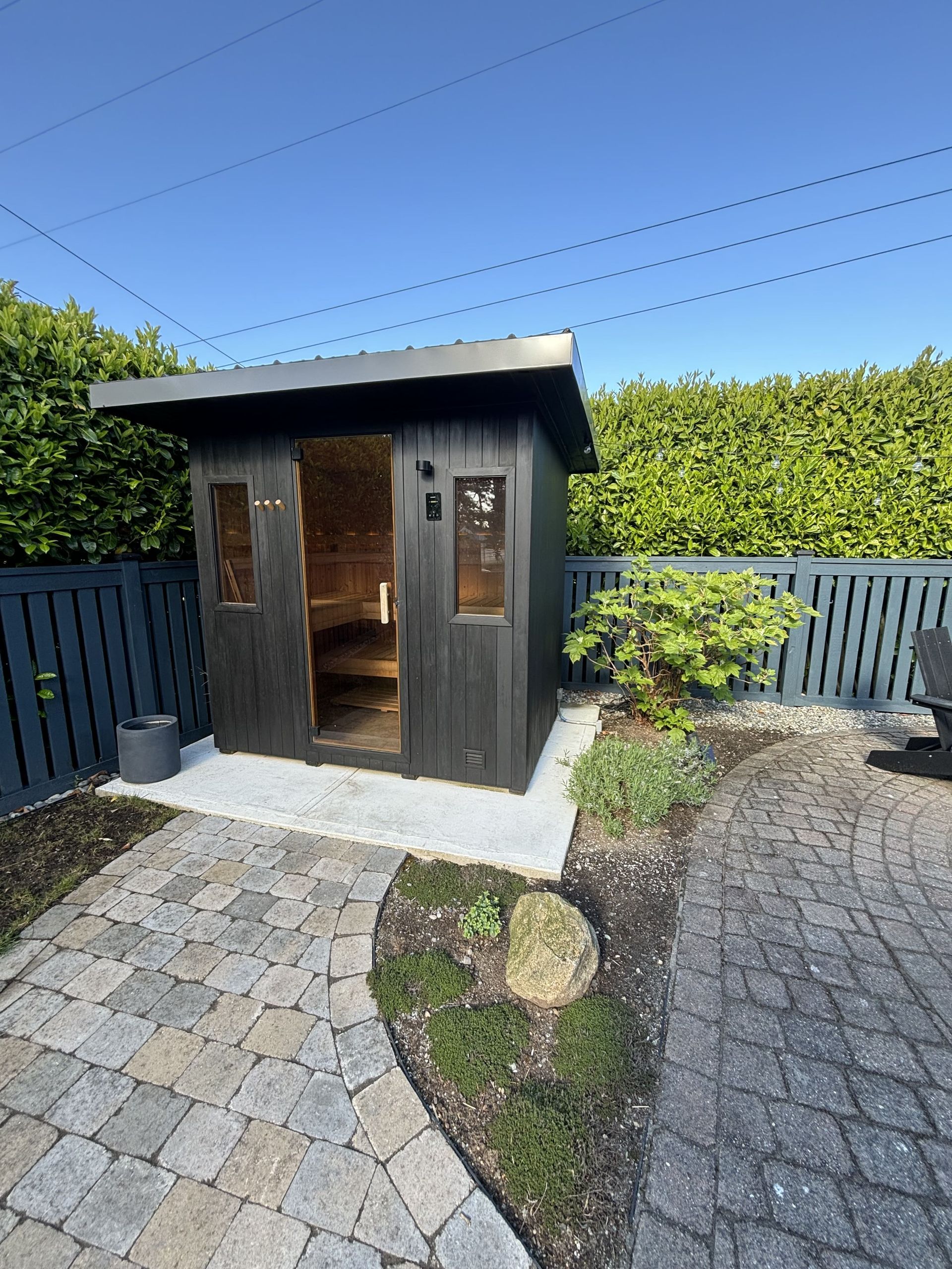 Small dark wooden garden shed beside a paved patio and hedge under a blue sky
