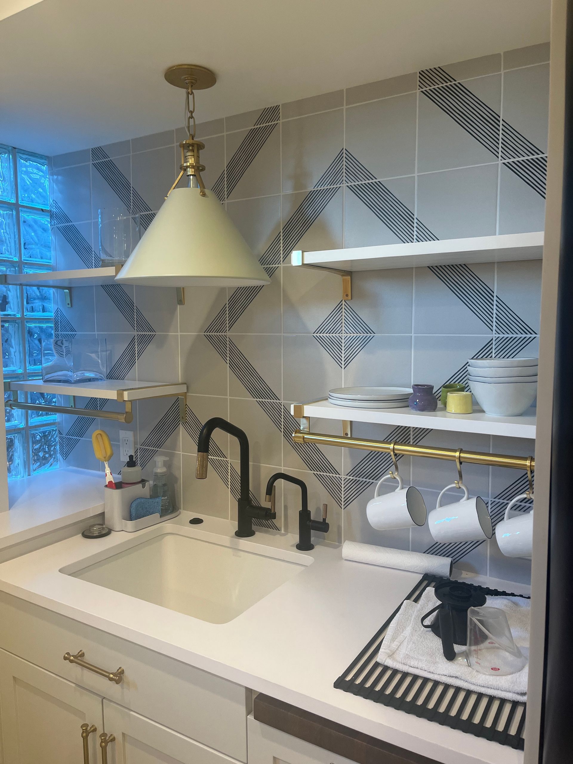 Modern kitchen with white sink, brass pendant light, patterned tile backsplash, and open shelves with dishes