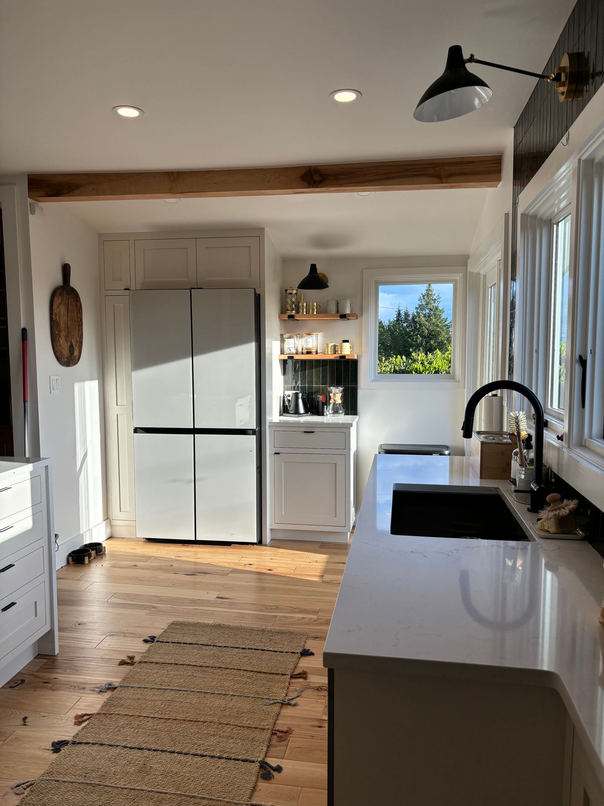 Bright farmhouse kitchen with white cabinets, black sink, wood floors, and a large fridge near a window.