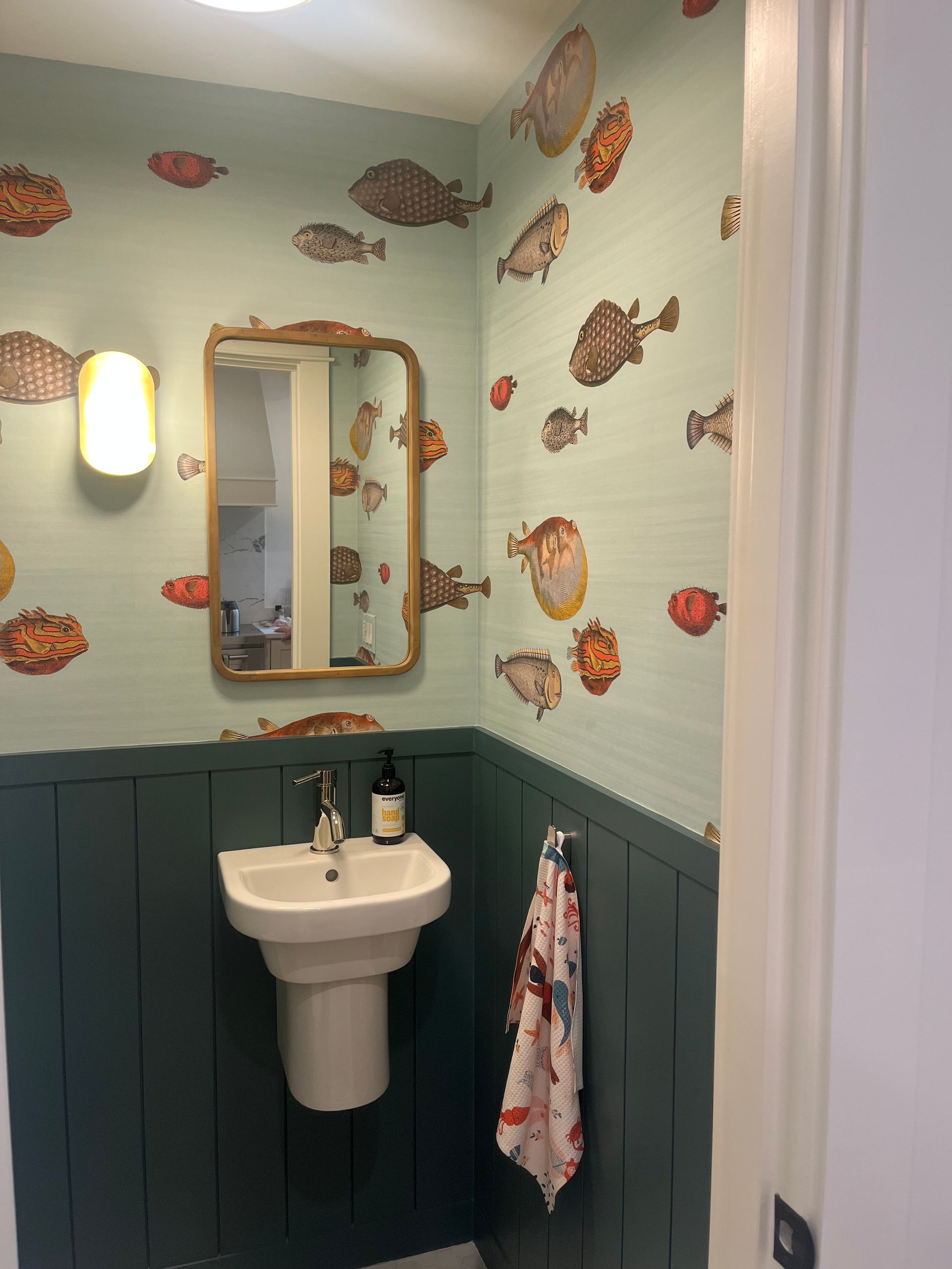 Small nautical-themed bathroom with fish wallpaper, sink, mirror, wall sconce, and hanging towel.