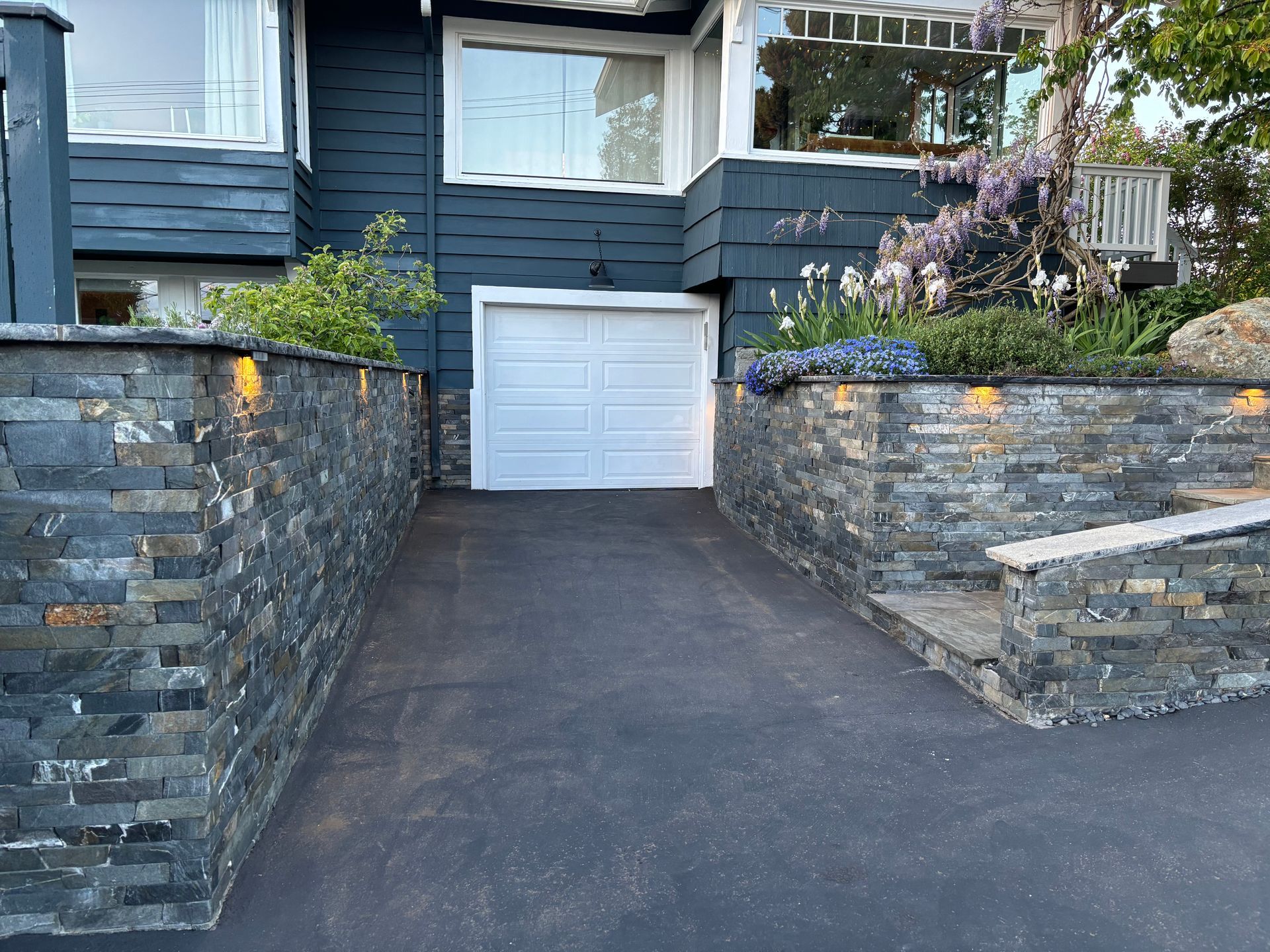 Stone driveway leading to a white garage door at a dark blue house with retaining walls and steps
