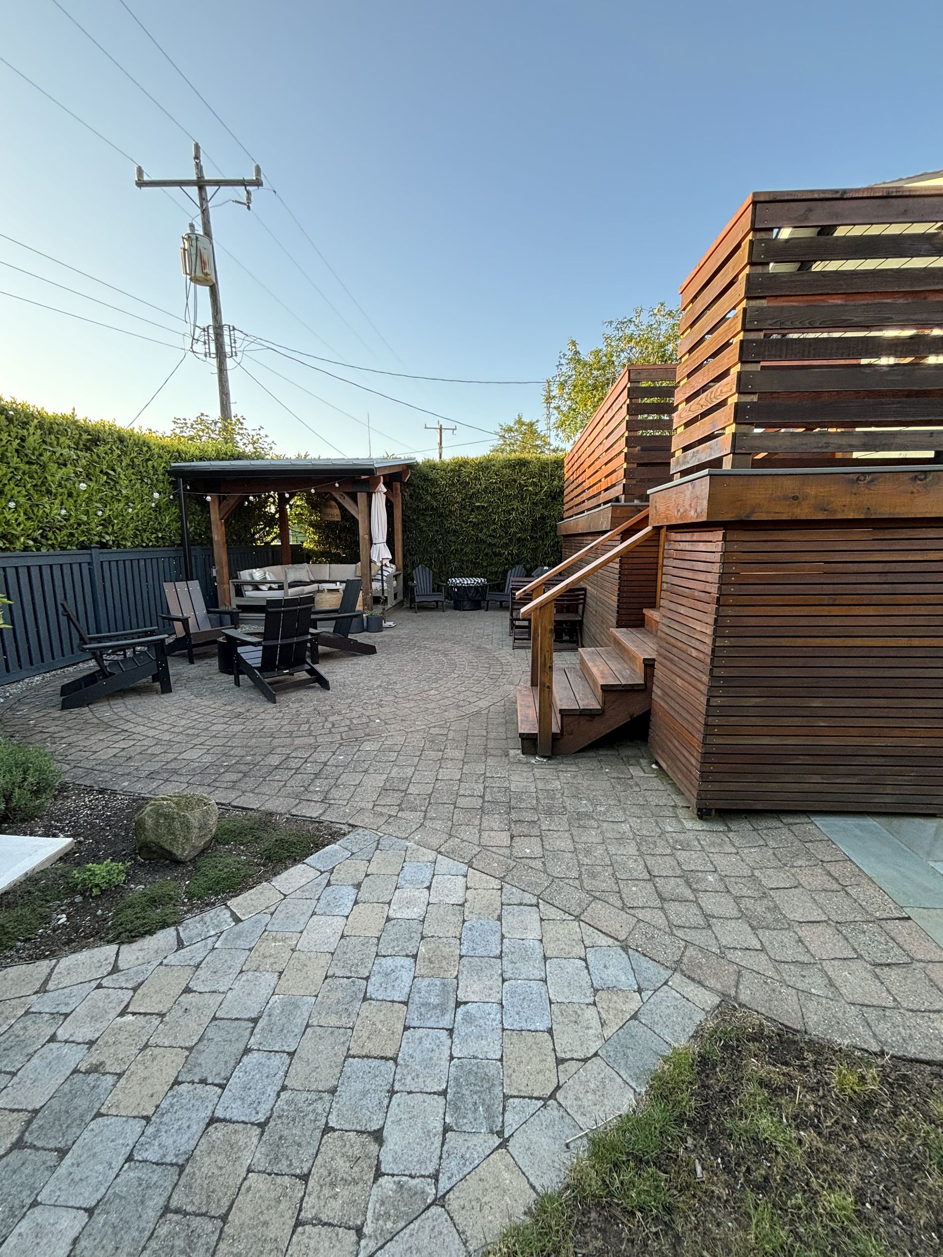 Backyard patio with stone pavers, wood pergola, hedge, and stacked lumber beside a gravel path