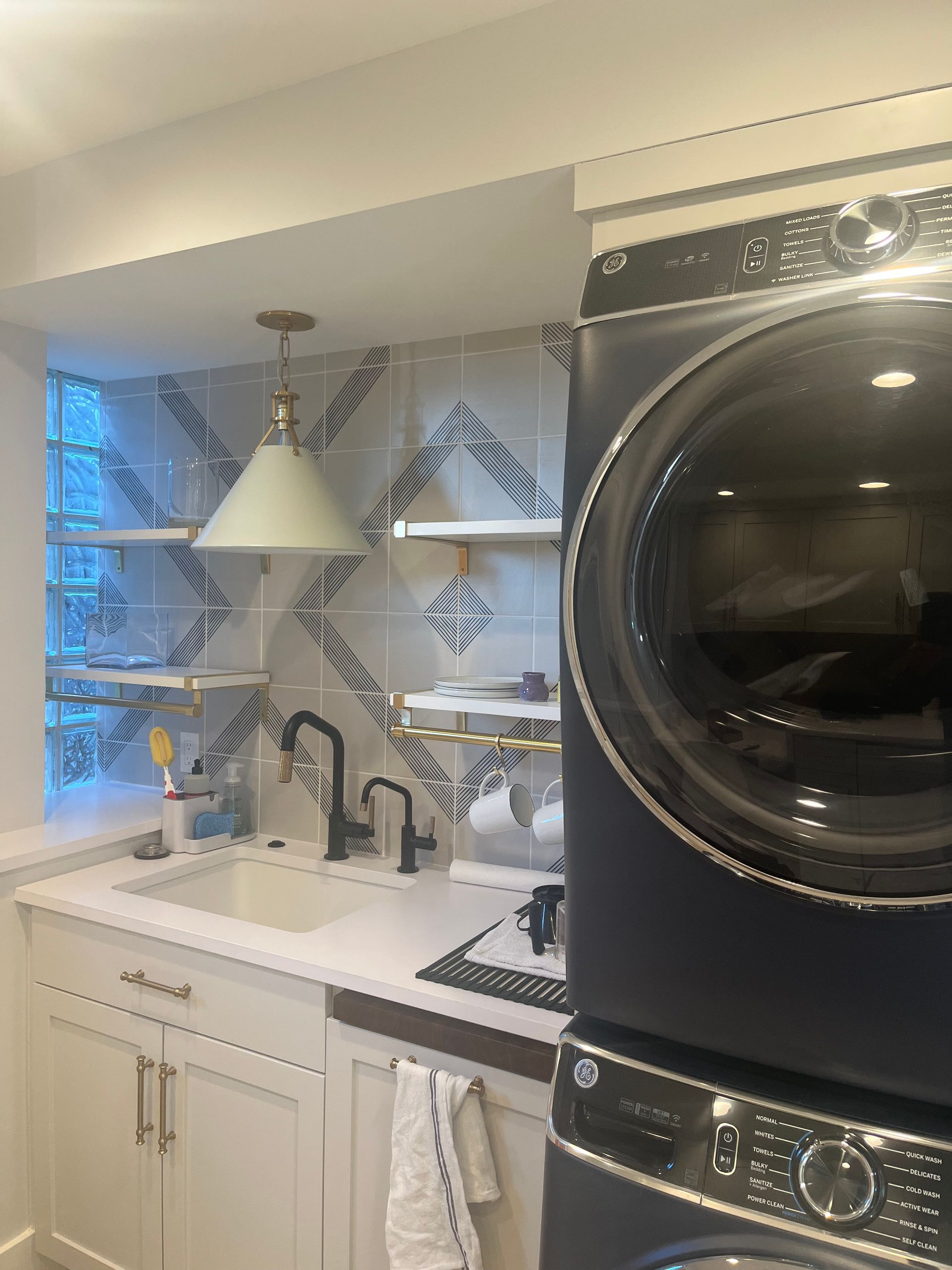 Compact kitchen with white sink, open shelving, pendant light, and stacked black washer and dryer.