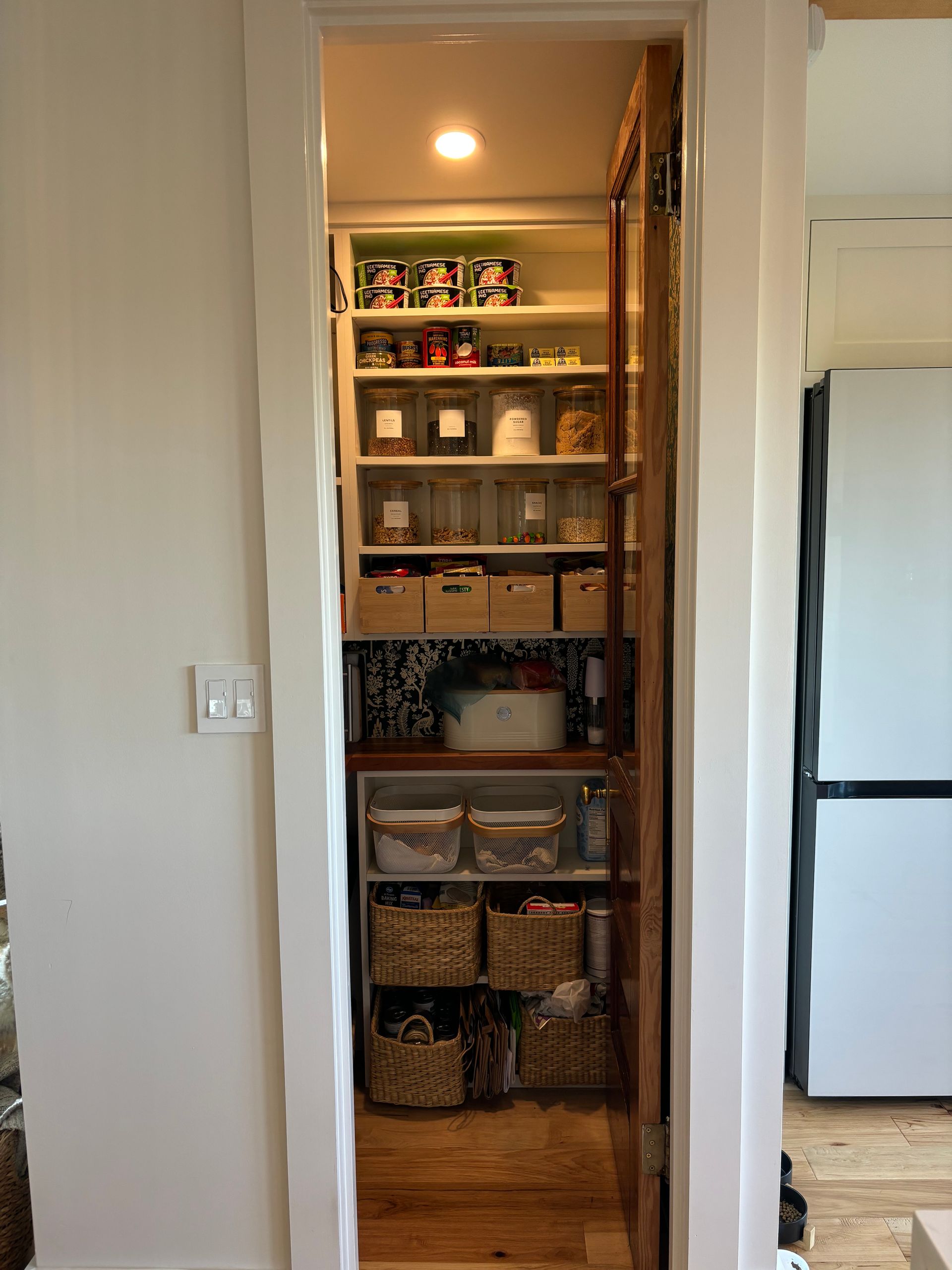 Open pantry closet with shelves of food, baskets, and storage bins in a hallway.