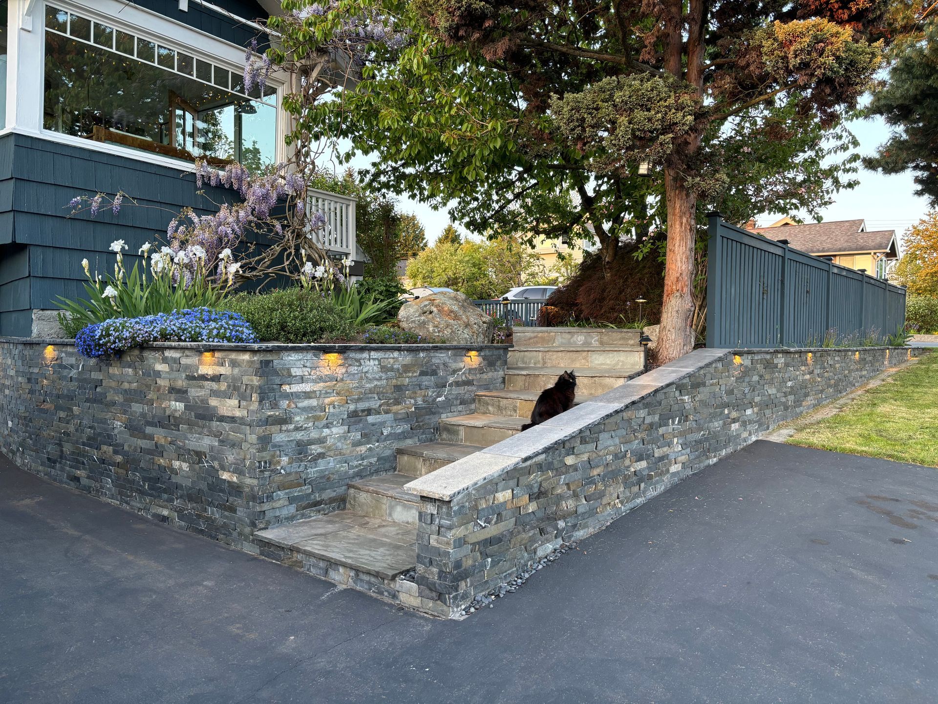 Stone retaining wall and ramp beside a house, with a black cat sitting near a tree and driveway.