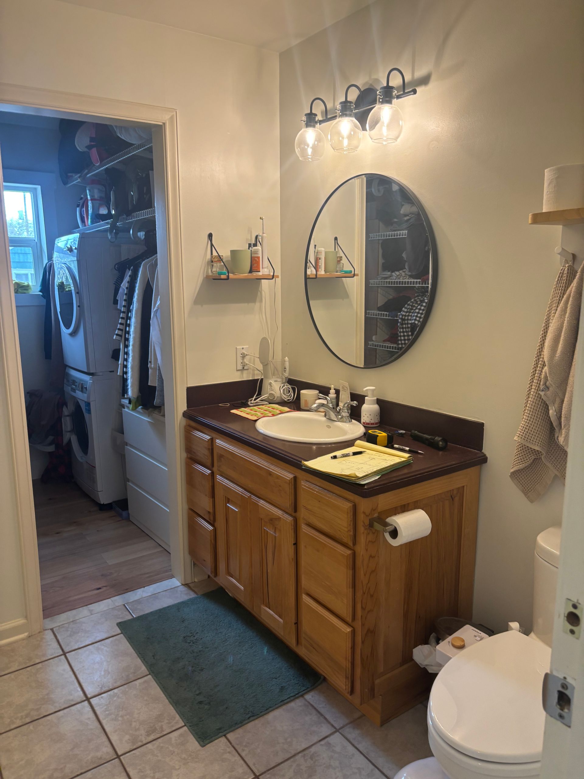 Bathroom with wooden vanity, round mirror, wall lights, tiled floor, and open doorway to a laundry area