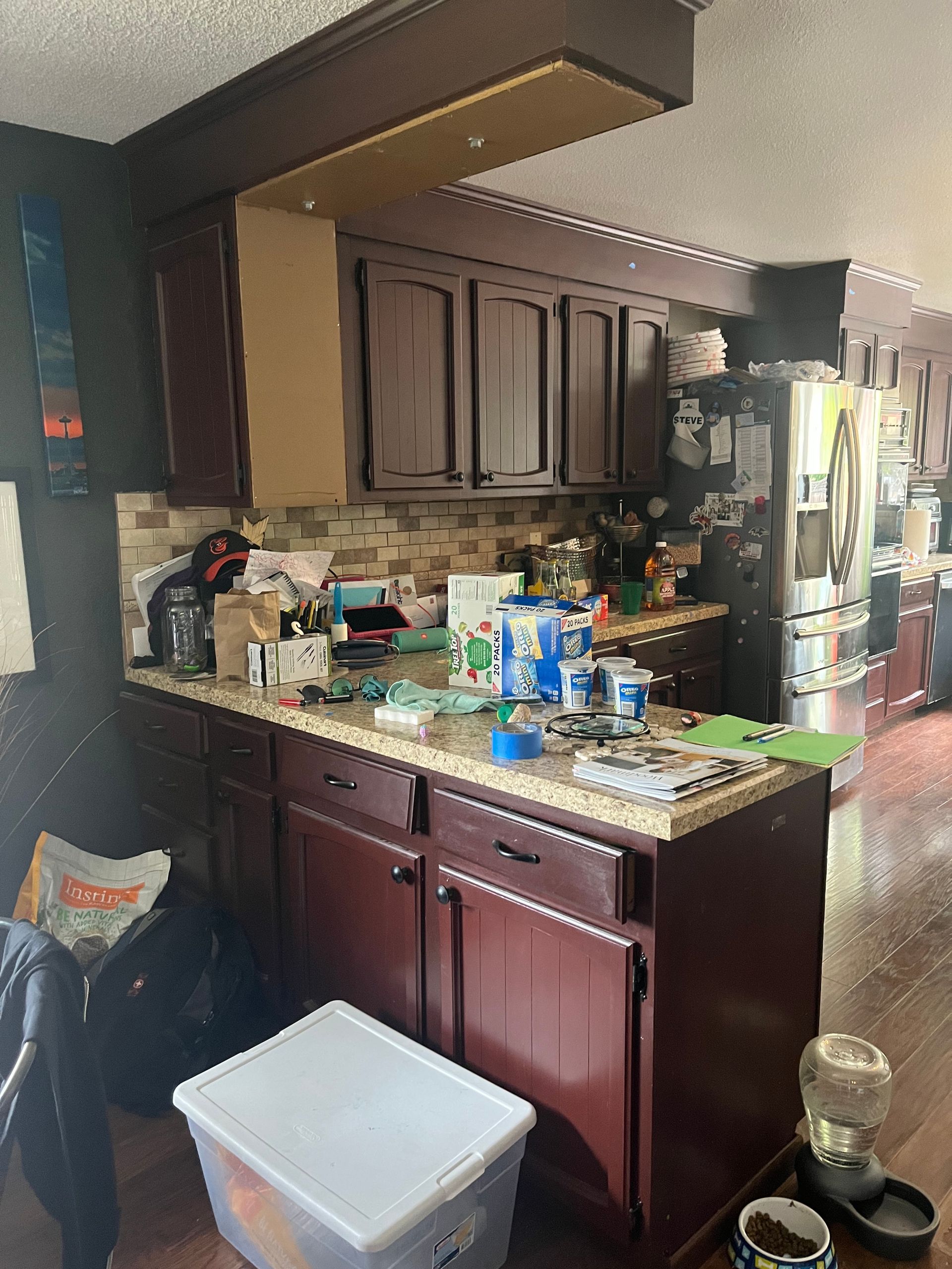 Cluttered kitchen with dark wood cabinets, countertop items, a refrigerator, and a white bin on the floor